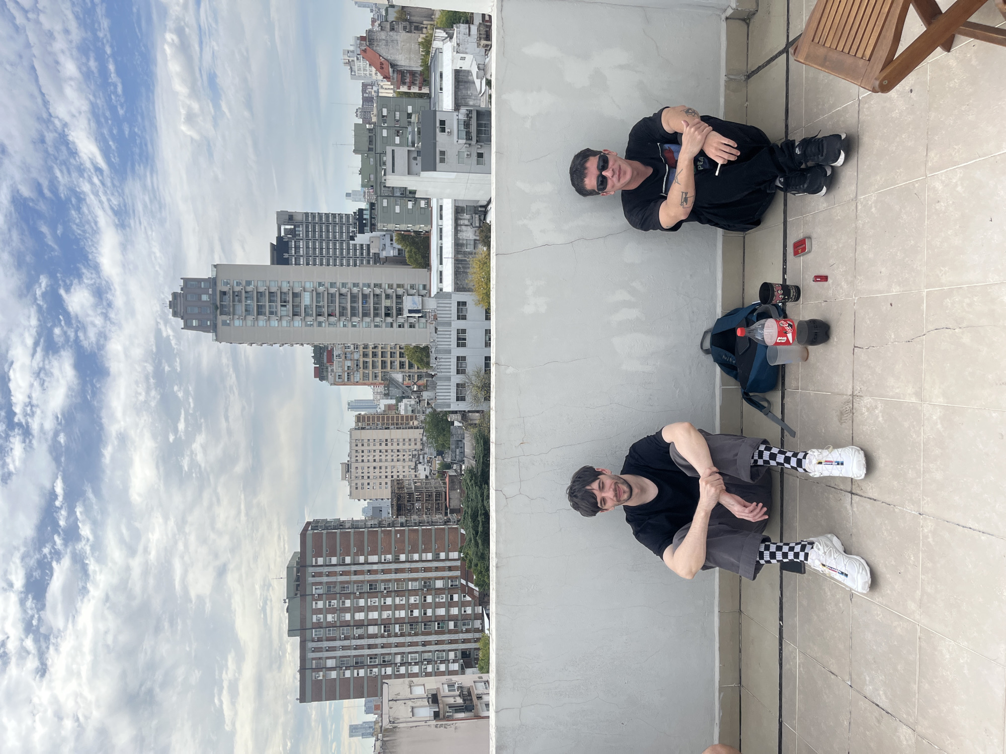 The image is a split-screen shot. On the left side, the view is of a city landscape with a cloudy sky. The buildings are in varying heights and styles, and include apartments and offices.
On the right side of the image, two men are seated on a rooftop or balcony. The first man, who is dressed in black, is seated at the top of the screen and is wearing sunglasses. He is holding a cigarette. The second man is seated at the bottom of the screen. He is wearing a black t-shirt and gray pants, and checkered socks and white shoes. Between them is a blue backpack, a red Coca-Cola bottle, cups, and a lighter. A wooden chair can also be seen at the top corner of the screen.
The buildings visible in the left side of the image indicate that the location is in Buenos Aires, Argentina.