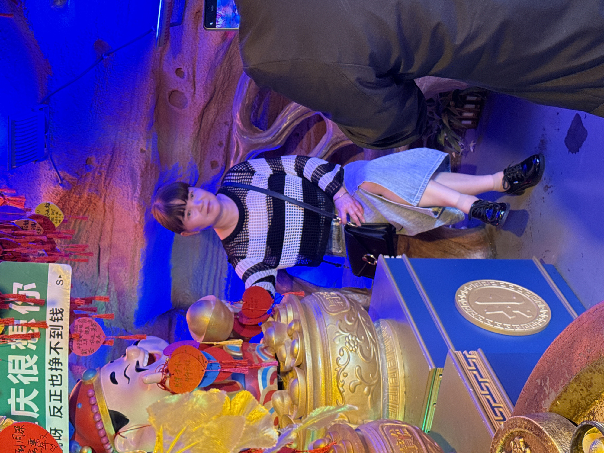 A woman is shown reclining sideways amidst an elaborate, illuminated display, with the location speculated to be in Yuzhong District, China. The display consists of a large, smiling figure with a festive hat and numerous decorative elements, including gold objects, red tags, and text in Chinese. She is wearing a striped sweater, a denim skirt, and black shoes and is holding a black purse. A person with dark pants is standing to the right. The background features textured surfaces illuminated with blue light.