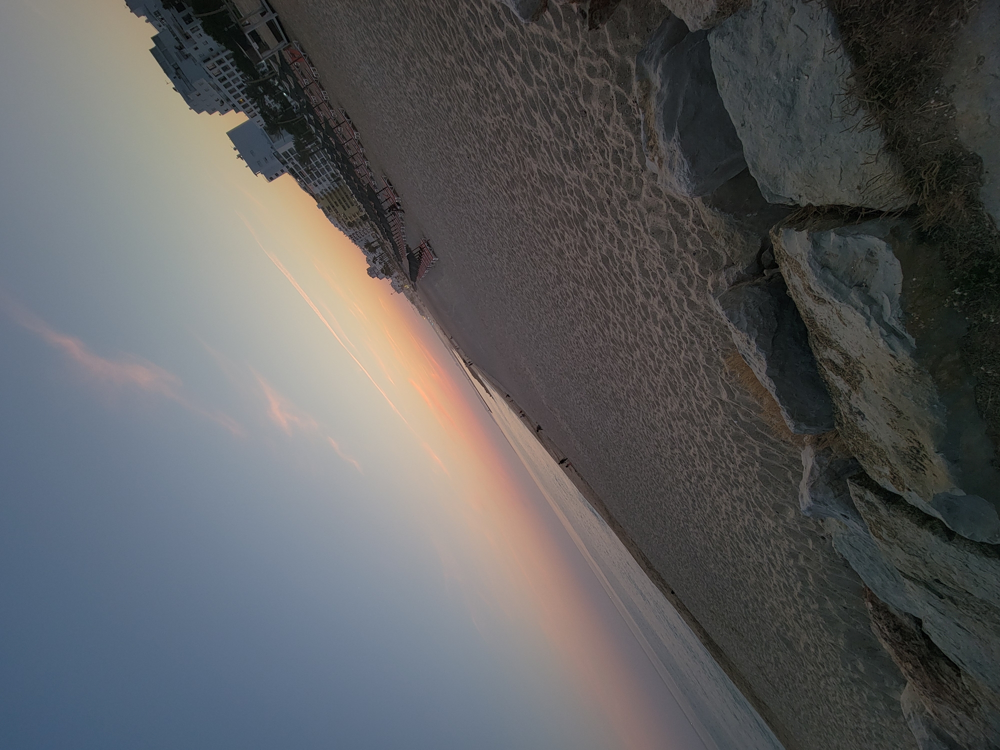 This image captures a scenic view of Quarteira, Portugal, at dusk. The sky transitions from a clear blue to soft hues of pink and orange, indicating the setting sun. A thin trail of clouds stretches across the upper part of the sky. The foreground features a sandy beach with scattered rocks, some of which are prominent in the lower right quadrant of the image. Beyond the beach, rows of empty beach chairs and umbrellas are arranged, suggesting a public beach. In the background, several multi-story white buildings, typical of a coastal resort town, line the shore. A few individuals can be seen walking along the beach, and the ocean is visible on the left, meeting the horizon. The perspective is slightly tilted, adding a dynamic feel to the composition.