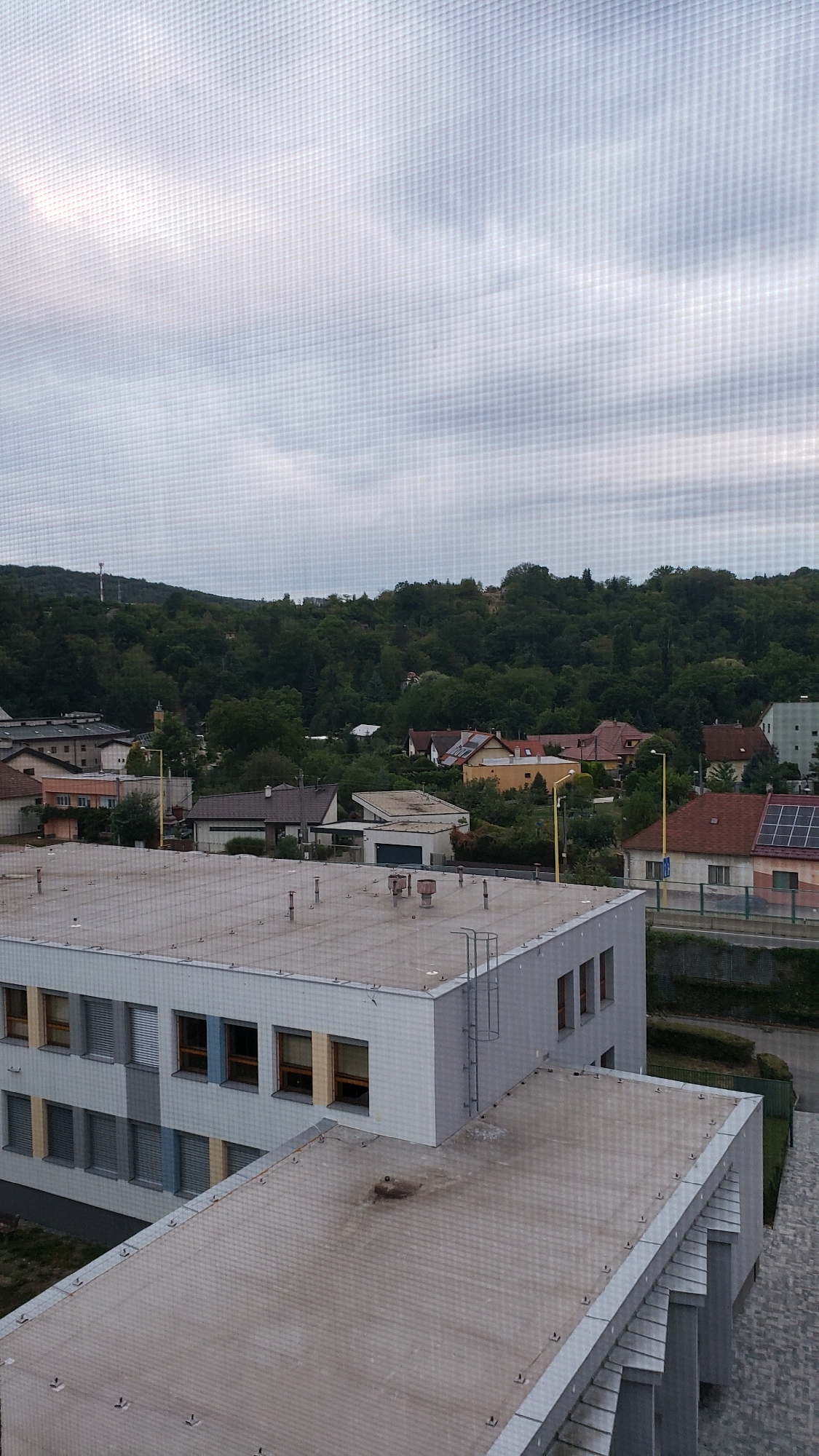 The image shows an aerial view of a suburban area. In the foreground, there is a large, flat-roofed building with several windows. Behind it, smaller houses with pitched roofs and some trees are visible. A densely forested hill forms the background. The sky is overcast with clouds. The image is taken through a mesh screen, which creates a grid pattern over the entire scene. The location is identified as Prešov, Slovakia.