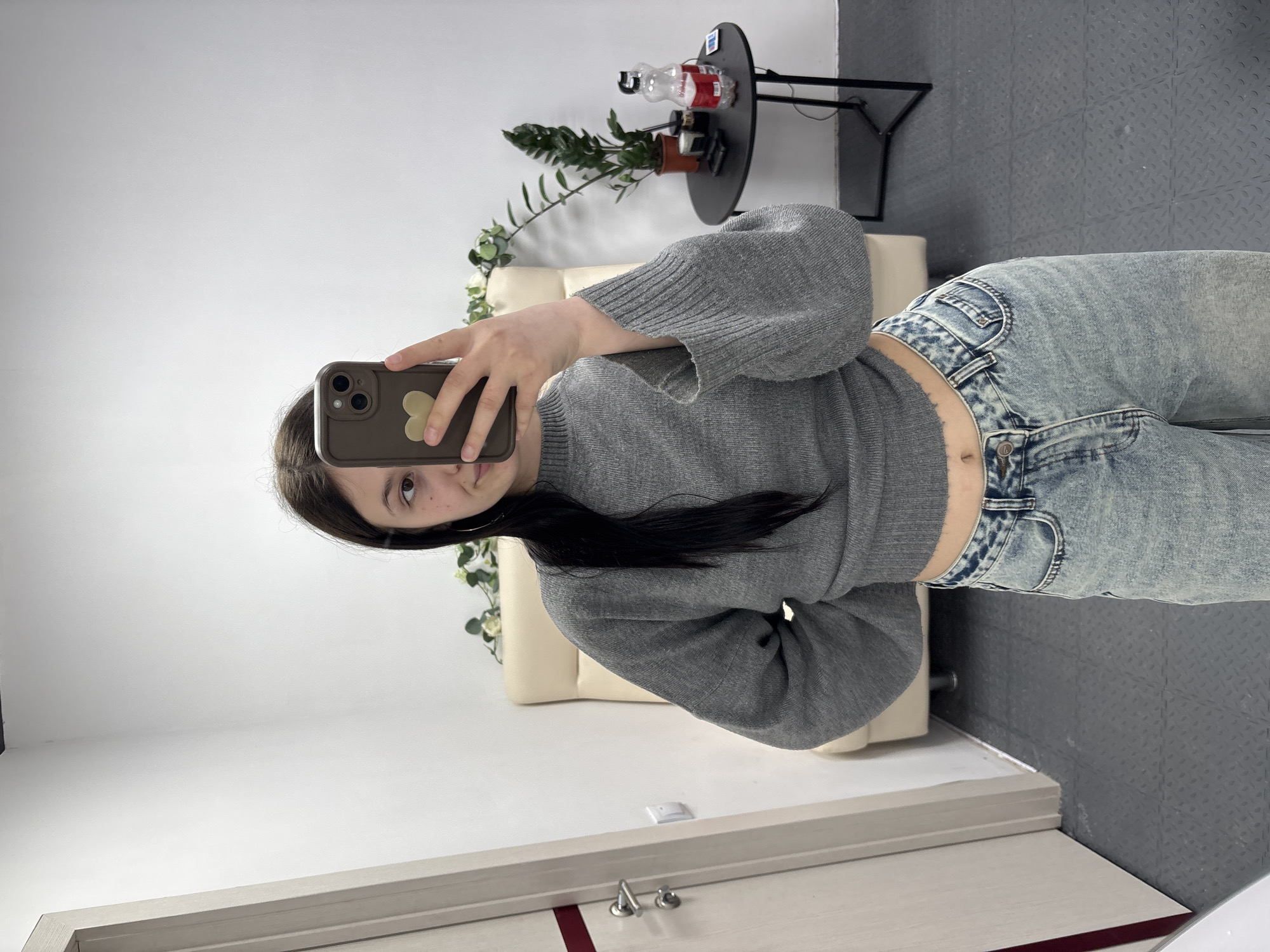 A young woman is depicted taking a mirror selfie indoors. She has long, dark hair and is wearing a gray knitted cropped sweater that reveals her midriff and navel, paired with light-wash denim jeans. She holds a smartphone with a dark case featuring a light-colored heart sticker, pointing it towards the mirror she is using (which is outside the frame). Her right eye and cheek are visible as she looks into the camera.
Behind her, a beige upholstered chair or bench is partially visible. To her upper right, a small round black table holds a potted plant with green leaves and two plastic bottles, one red and one clear. The room features plain white walls and a dark gray floor with a distinct textured pattern, possibly a diamond plate or studded design. A light-colored door frame with silver-colored hardware and a red strip is visible in the foreground. The setting suggests a casual indoor space, possibly in Фоки, Russia. The lighting is internal, so the time of day is not indicated. There is no visible text in the image.