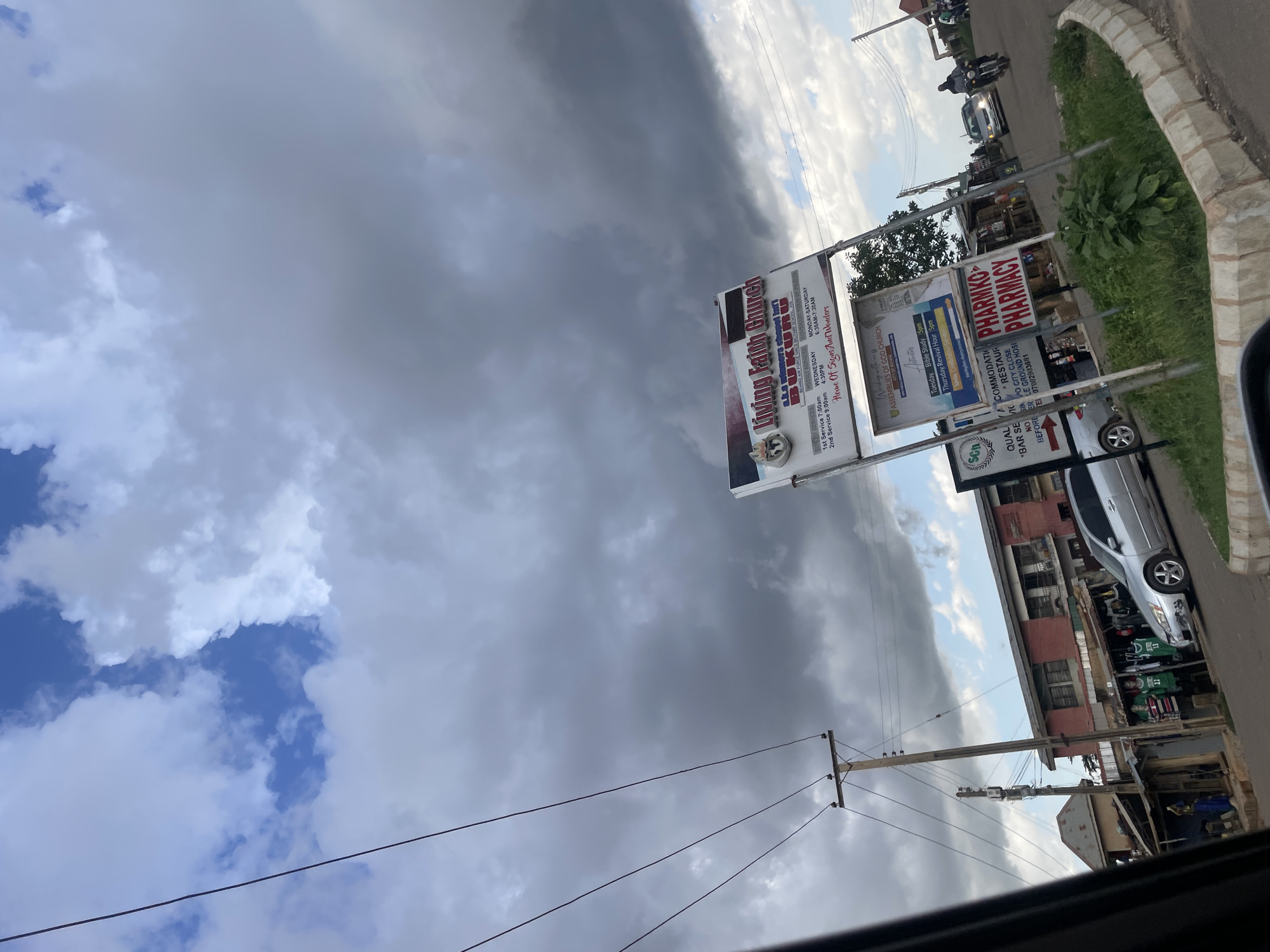 The image depicts a street scene in Gura Top, Nigeria, under a dramatic sky filled with heavy, grey clouds and patches of blue. A prominent sign for "Living Faith Church Bukuru" stands on the right side of the frame, accompanied by smaller signs for "Assemblies of God Church" and "Pharmko Pharmacy." The church sign details service times and locations. In the mid-ground, a silver sedan is parked on the street, and to the left, a row of buildings houses market stalls with clothing and merchandise displayed. Power lines and utility poles are visible throughout the scene, suggesting an urban or semi-urban environment. A few individuals are present, including people near the market stalls and figures on a motorcycle further down the road, indicating ongoing daily activity. The overall atmosphere is overcast, with the clouds suggesting potential rain.