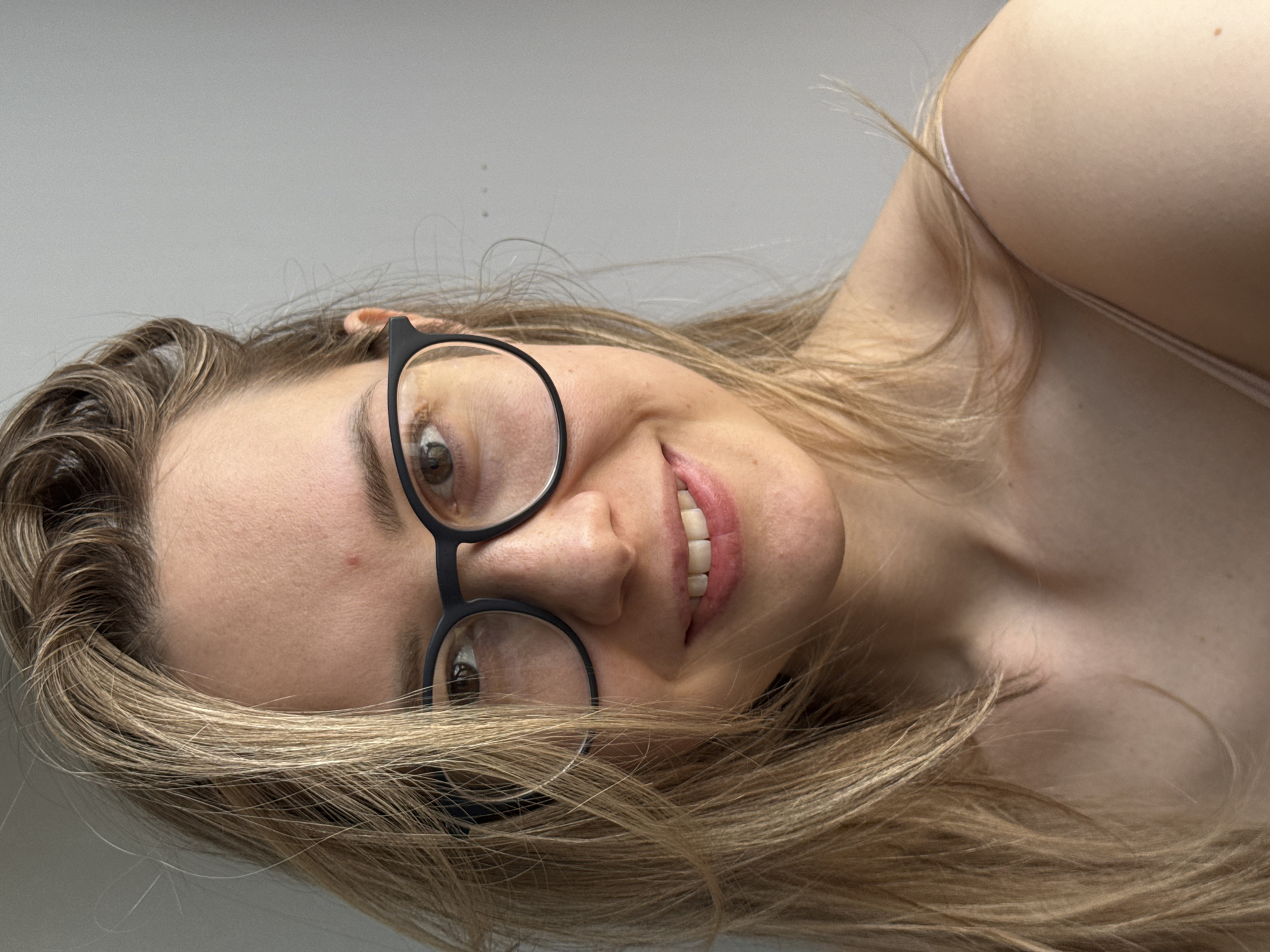 A close-up portrait shows a young woman smiling and looking directly at the viewer. The image is rotated counter-clockwise by approximately 90 degrees. She has long, light brown to blonde hair that frames her face and flows around her. She is wearing black-framed eyeglasses, revealing her brown eyes. Her lips are parted in a wide, natural smile, showing her white teeth. Her skin has a fair complexion, and a small blemish or mole is visible on her forehead.
She appears to be wearing a light-colored top, with a thin strap visible over her right shoulder, suggesting a tank top or camisole. The background is a plain, light gray or off-white surface, indicating an indoor setting. The lighting is bright and even, highlighting her features. Her expression is pleasant and friendly. There is no visible text in the image.