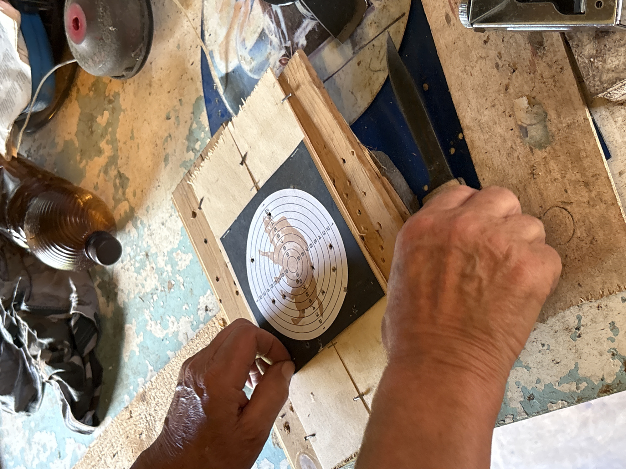 A close-up, overhead shot shows a person's hands working on a wooden board with a target attached, placed on a weathered blue and white painted surface. In the foreground, two hands are visible. The left hand is positioned to the left of a circular target, and the right hand is to the right of the target, holding a knife blade. The target, a black square with a white circular bullseye marked with concentric rings and numbers, is affixed to a piece of plywood. Small nails are used to attach the plywood to another wooden surface underneath. In the background, a large plastic bottle with amber liquid and a dark cap is visible, along with a crumpled piece of fabric and a red and gray object with a red center. The overall setting appears to be an outdoor or workshop environment. The image is taken in Дресвяново, Russia.