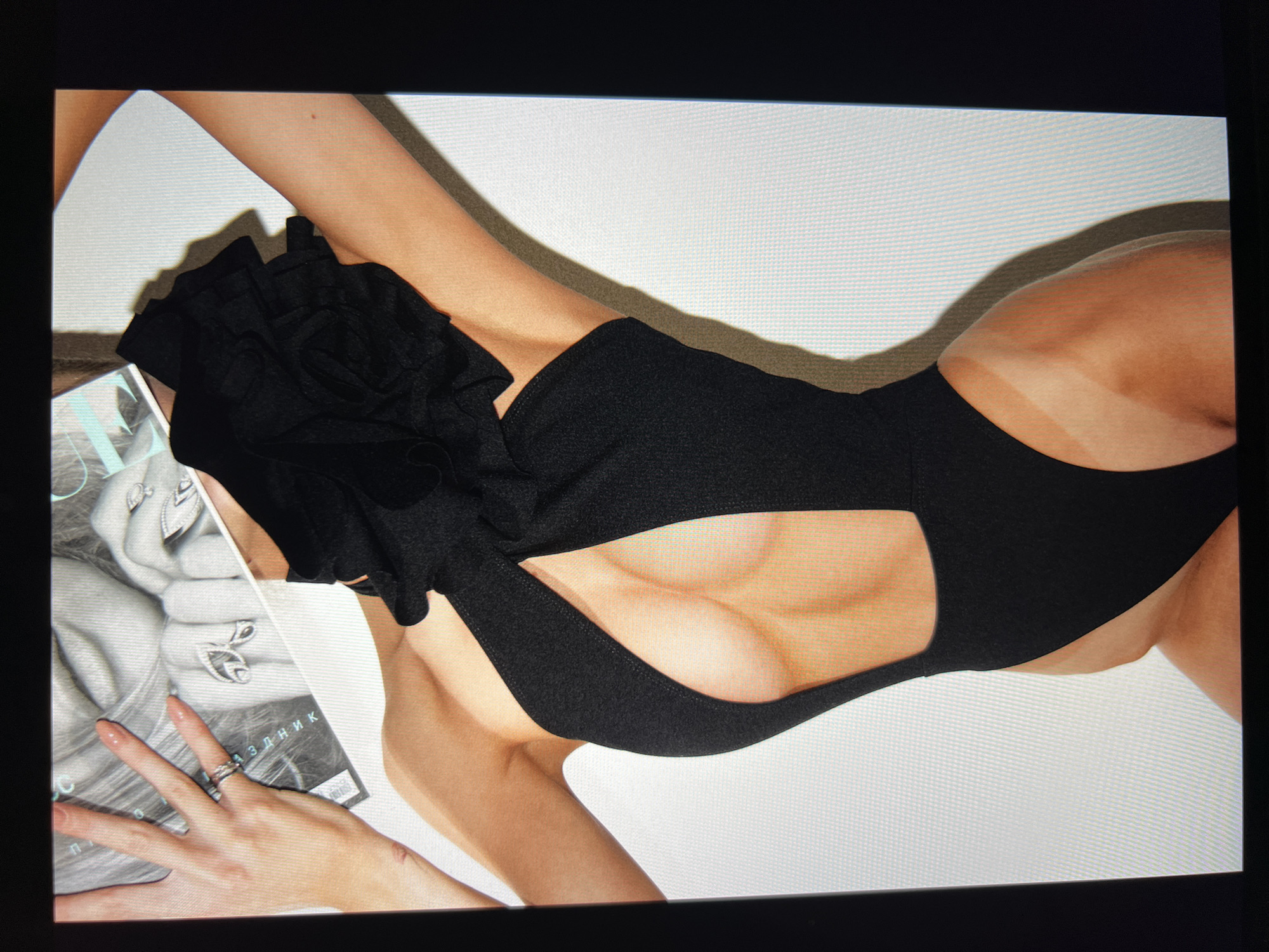 In an indoor setting with a plain white wall, a person is positioned on their side, their body facing left. They are wearing a black one-piece swimsuit with a cutout on the chest and a large floral embellishment. The swimsuit has a halter neck and straps that go over the shoulders. The person's left arm is extended upwards, holding a magazine or book. Their right arm is bent, with their hand resting on their chest. There is a visible tan line on the person's hip and thigh. The lighting creates a shadow on the wall behind them, indicating a light source to the right. The scene is captured from a medium shot, focusing on the upper body and part of the legs.