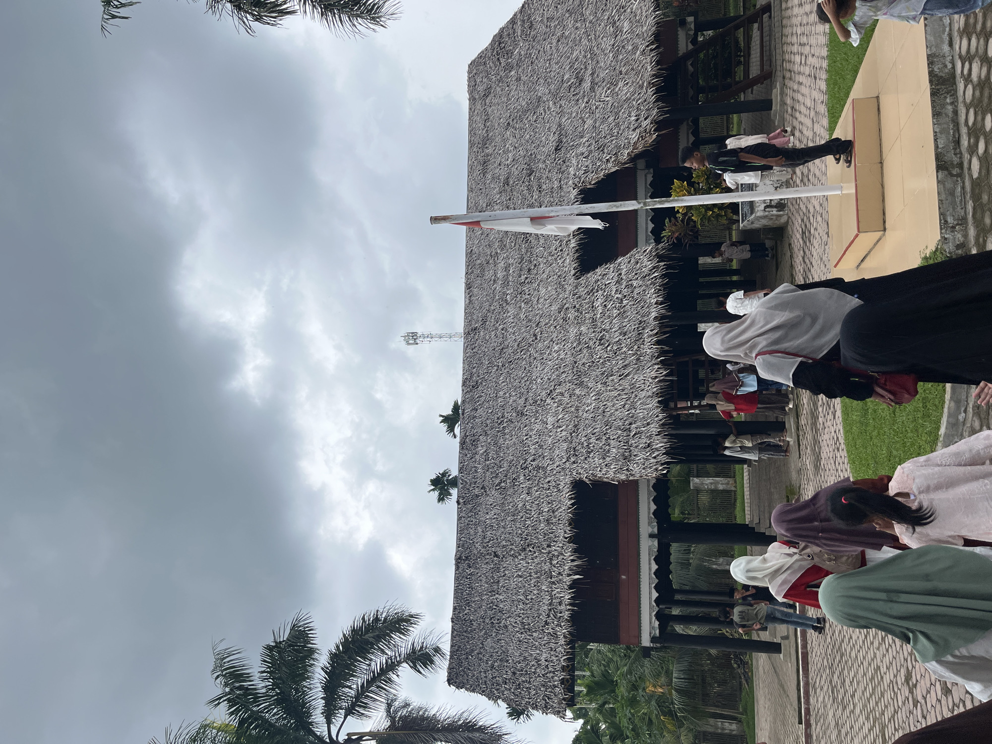 A group of people, many wearing hijabs, walk on a stone pathway towards a traditional Indonesian house with a thatched roof. A flag flies from a pole in front of the house. In the background, palm trees and other greenery are visible under a cloudy sky. A cell tower is also visible in the distance. The scene appears to be an outdoor cultural site or historical landmark.