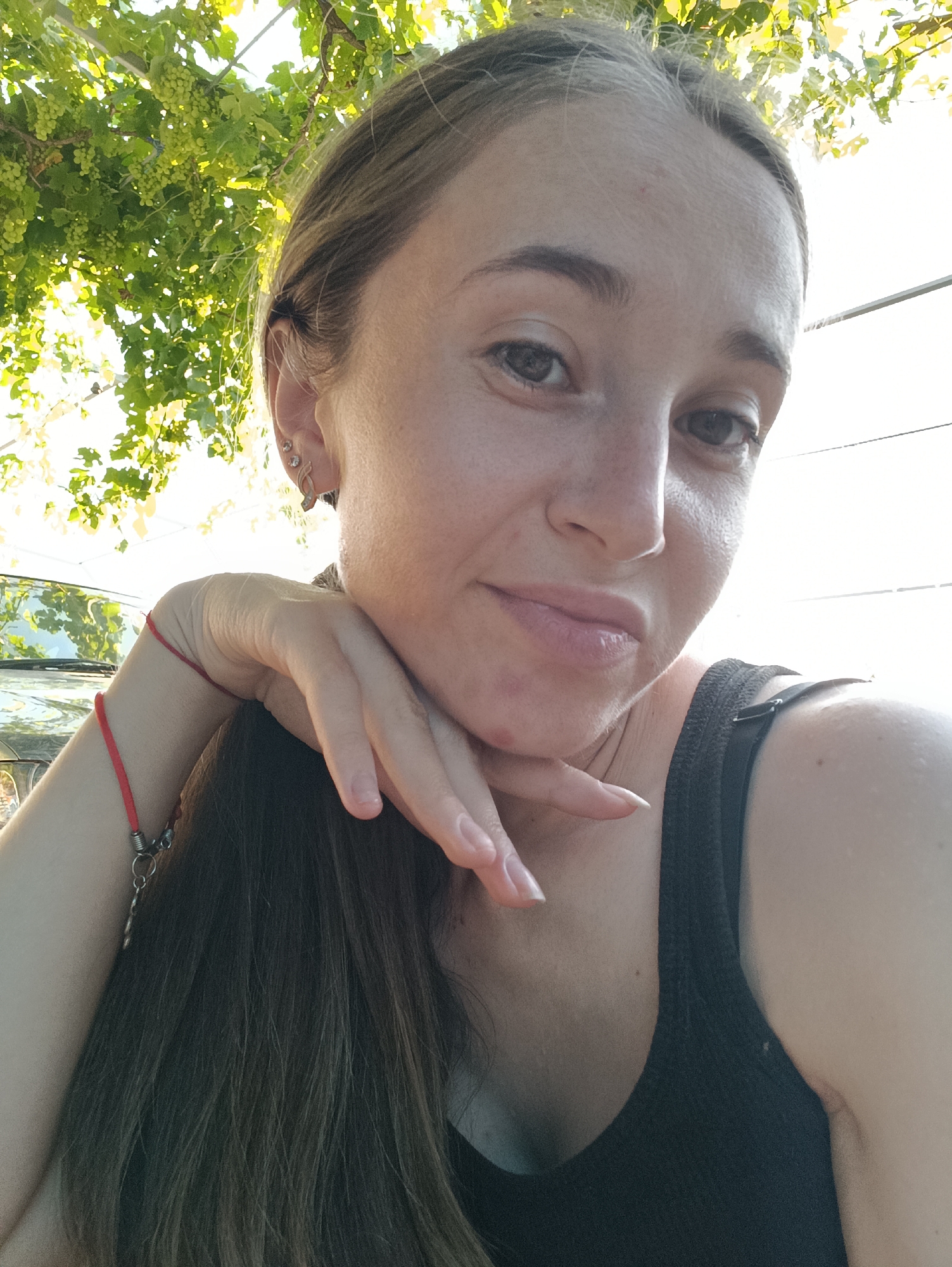 A young woman, with light brown hair and fair skin, is taking a selfie outdoors. She is wearing a black tank top and a red bracelet on her left wrist. Her hand is resting on her chin, and she is looking directly at the camera with a slight smile. In the background, there are green grapevines with clusters of grapes, and sunlight filters through the leaves, creating a dappled effect. A portion of a dark-colored vehicle is visible to the left of the woman. The scene appears to be set in a garden or vineyard, possibly in the city of Vypasne, Ukraine, given the mention of grapes.