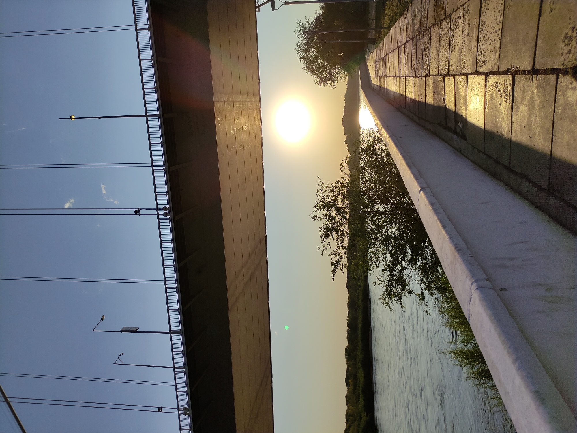 The image shows a view from under a bridge in the city of Semey, Kazakhstan, overlooking the Irtysh River. The sun is setting, casting a warm glow on the scene. A paved walkway runs alongside the river, bordered by a white wall. Trees and vegetation line the opposite bank of the river. The bridge above is supported by cables, and lights can be seen on its deck. The sky is clear with a few wispy clouds. The scene conveys a sense of tranquility and natural beauty.