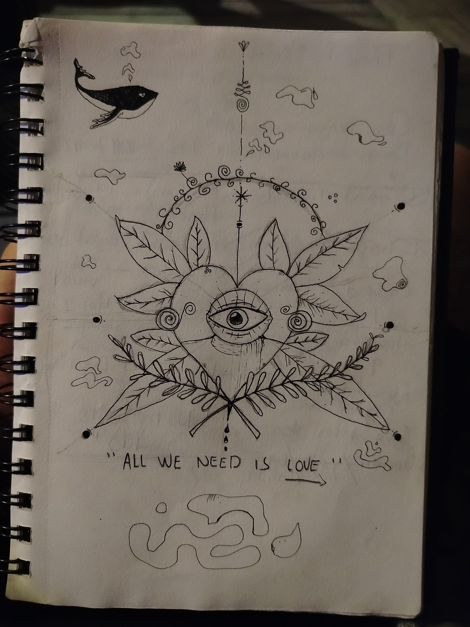 A black and white sketch on a spiral-bound notebook depicts a stylized image with a central heart-shaped form containing a single eye. The heart is surrounded by leaves and swirling decorative elements. Above the heart is a vertical line with symbols and circles. At the top left is a drawing of a whale, emitting bubbles. Scattered around the central image are cloud-like shapes. Below the heart is the text "ALL WE NEED IS LOVE" with an arrow pointing to the right. Underneath the text are abstract, amorphous shapes. There is no indication that the image is in Playa del Carmen, Mexico.