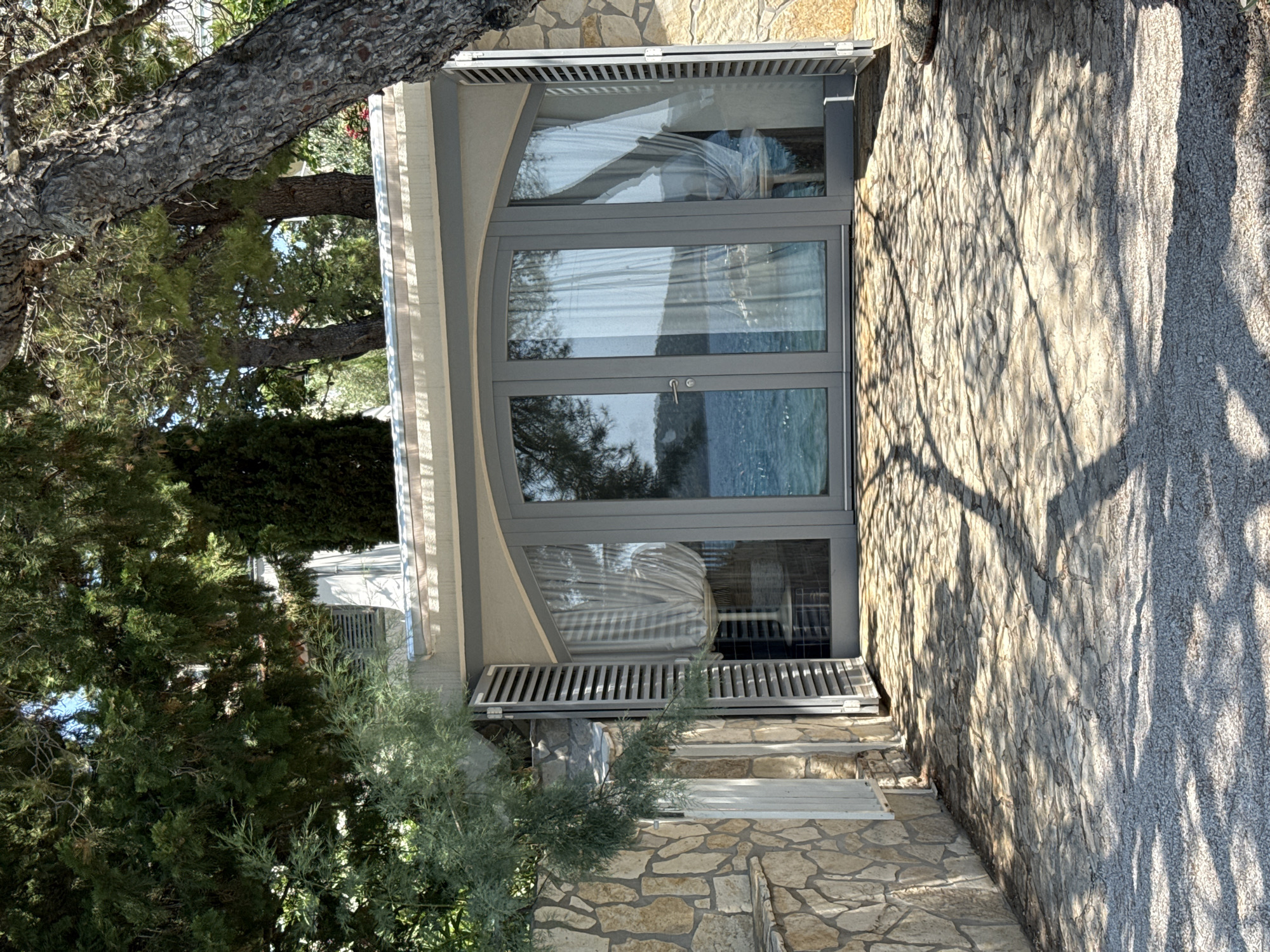 This is an eye-level, medium shot of a modern building with large glass doors facing a body of water, likely the Adriatic Sea, in Grad Šibenik, Croatia. The building's exterior is made of light-colored stone, and it is partially obscured by lush green trees. The foreground is a stone patio with a gravel border, and tree shadows are cast across the patio. The glass doors are framed by gray shutters, one of which is open. Inside the building, sheer white curtains are visible. The scene appears to be a residential or commercial property, possibly a vacation rental, given its waterfront location and modern design.