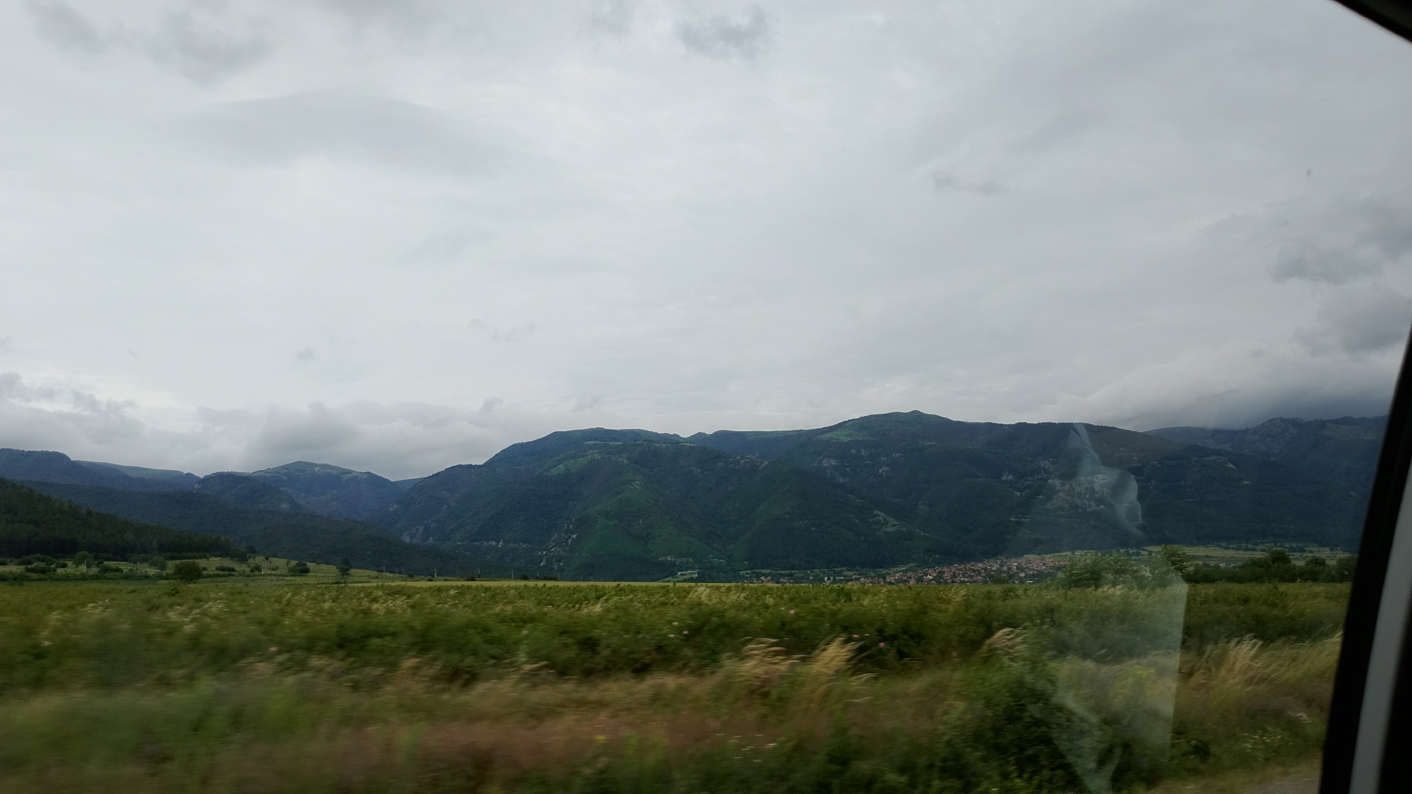 The image shows a view from inside a vehicle, looking out at a rural landscape. In the foreground, there is a blurred field of tall green grass and some dry, straw-like vegetation. Beyond the field, a range of dark green, tree-covered mountains dominates the middle ground. The mountains are vast and rolling, with visible valleys and slopes. In the far distance, a small town or village with red-tiled roofs is nestled at the base of the mountains. The sky is overcast with thick, gray clouds, suggesting a cloudy or possibly rainy day. A dark frame on the right edge of the image indicates the presence of a car window or similar enclosure. There are no people visible in the scene.
