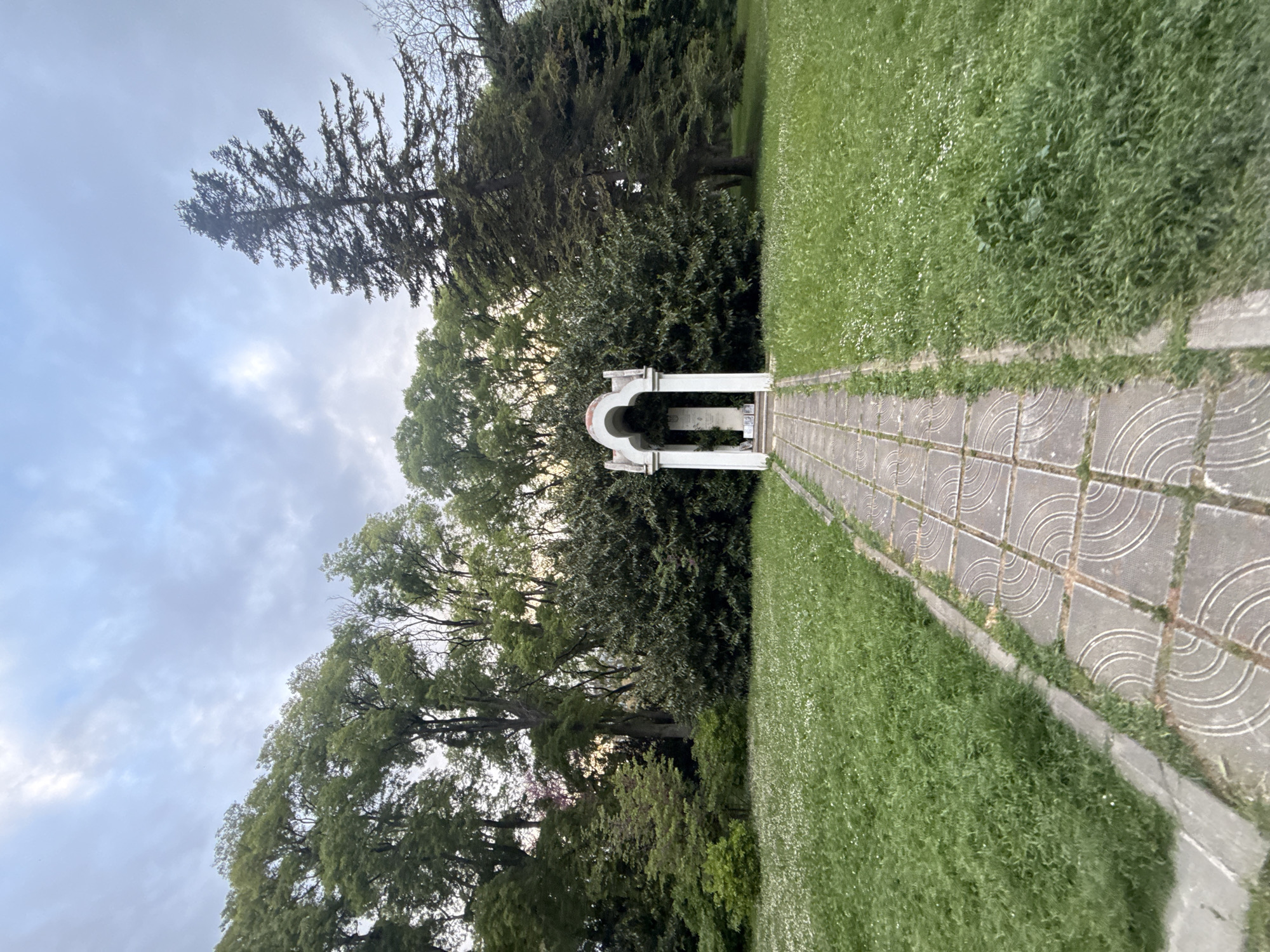 The image shows a pathway made of square concrete pavers leading to a small white arched structure. The pathway is bordered by green grass on either side. The structure is surrounded by trees and shrubs. The sky is cloudy. The photo was taken at Primorski park in Varna, Bulgaria.