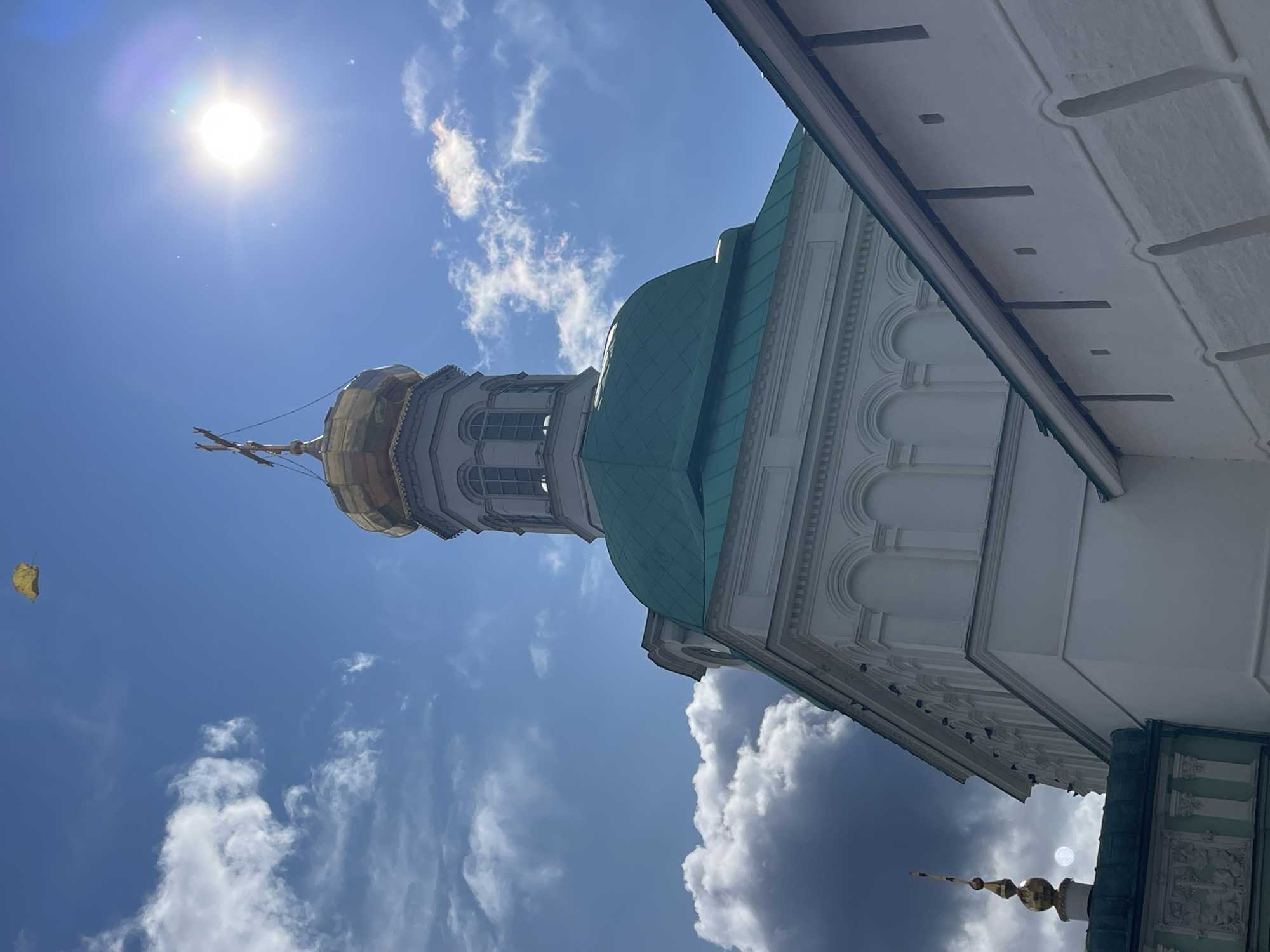 The image captures the Trinity Lavra of St. Sergius in Sergiyev Posad, Russia, on a bright, sunny day with scattered clouds. A golden dome topped with a cross is visible on a white church structure. The dome is part of the assumption Cathedral. Adjacent to this is a green-tiled roof and a white wall. The sun is high in the sky, creating a lens flare. A single yellow leaf is seen falling in the upper left corner of the sky. The scene is focused on the architectural details of the monastery and the sky above.