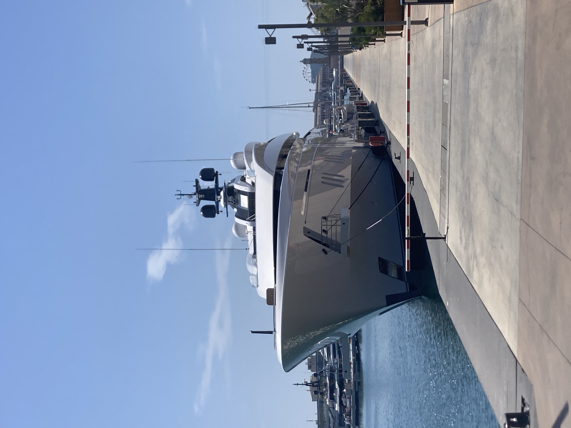 This media file captures a scene at a marina, likely in Barcelona, Spain, given the context provided. The dominant subject is a large, modern white yacht, prominently docked along a pier. The yacht is sleek and luxurious, with multiple decks and a prominent radar or communication array at its top. Its hull is a clean white, contrasting with the dark blue water it floats on.
The setting is a sunny day, indicated by the bright blue sky with a few wispy clouds and the sharp, clear shadows cast by the objects. The time of day appears to be late morning or early afternoon due to the angle of the sun. The pier is made of concrete and runs alongside the yacht, with a red and white striped barrier extending from a post. In the background, other boats are visible docked along the marina, suggesting a bustling maritime environment. Further beyond the immediate marina, there are buildings and a Ferris wheel, hinting at a more urban or resort-like area adjacent to the harbor.
No people are clearly visible in the image, focusing the attention on the impressive vessel and its surroundings. The overall mood conveyed is one of calm and luxury, characteristic of a high-end marina. The environment is clean and well-maintained, with the water appearing calm and reflecting the bright daylight. No visible text is discernible within the image itself.