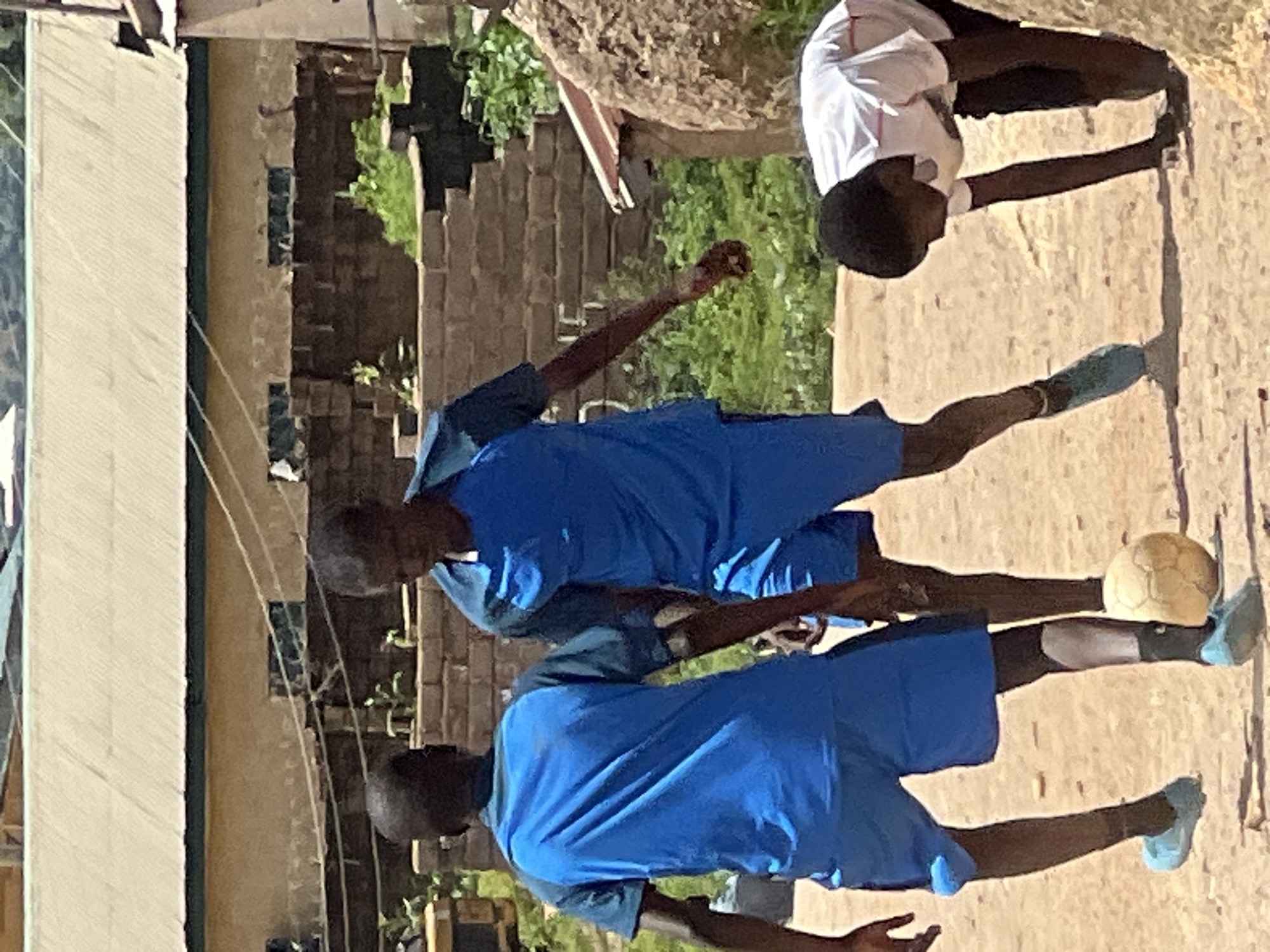 In Jos, Nigeria, three young boys are engaged in a game of soccer. Two boys, dressed in matching blue soccer uniforms, are actively participating in the game. One boy, facing away from the camera, is positioned to the left, while the other, facing the camera, is in the center. A third boy, wearing a white t-shirt and dark shorts, is bending down to the right, possibly retrieving the ball. The ground is dusty and appears to be a dirt field. In the background, there is a building with a corrugated metal roof and brick walls, suggesting a residential or communal area within the city. The scene conveys a sense of casual recreation in an urban Nigerian setting.