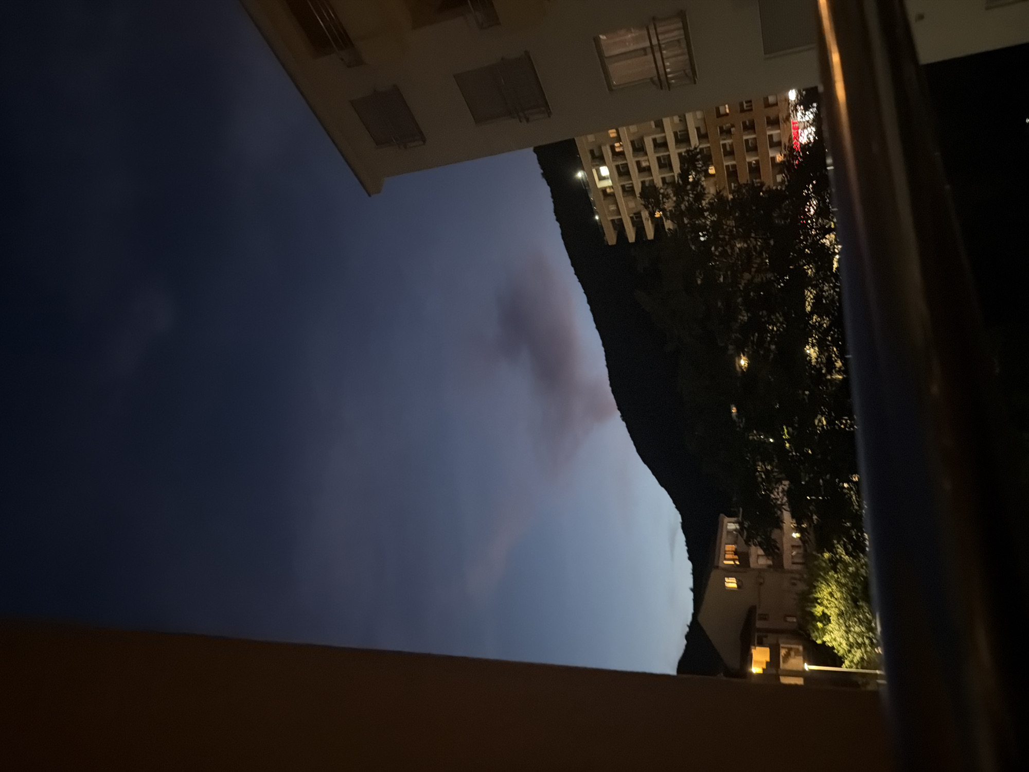 The image shows a view from a balcony at dusk, looking towards a mountainous landscape with buildings. The sky is a deep blue, transitioning to lighter shades near the horizon, with some wispy clouds. A faint plume of smoke or dust is visible rising from the mountainside. On the right, a modern, multi-story apartment building with white exterior walls and balconies is prominent. To the left and center, other buildings are visible, some with illuminated windows, suggesting they are residential or commercial. Trees and greenery are interspersed between the buildings and the mountains. The foreground shows a dark railing, likely part of the balcony from which the photo was taken. The scene captures a moment of twilight in a city, possibly Budva, Montenegro, given the mountainous terrain and architectural style of the buildings. There are no people or specific actions depicted.