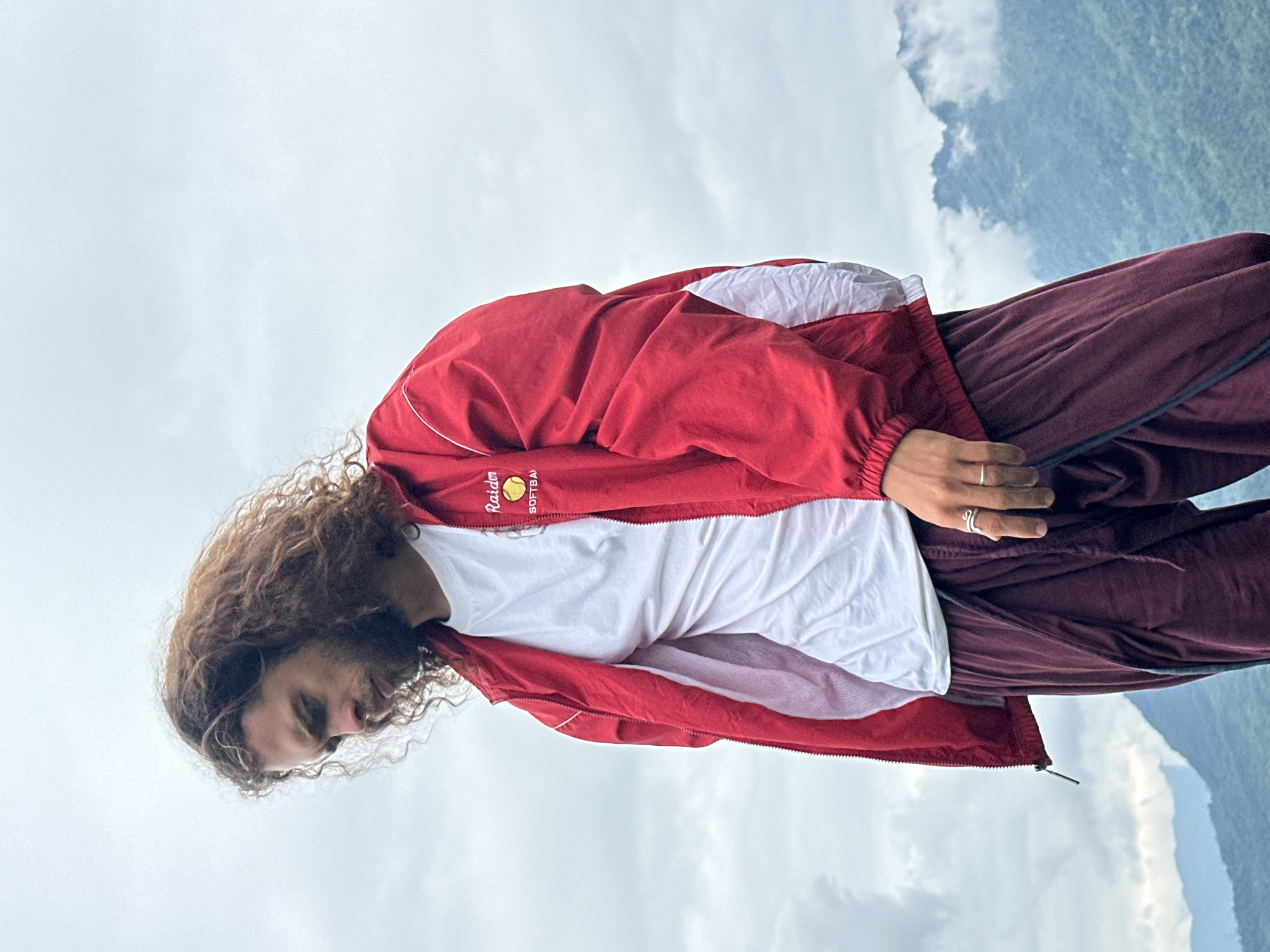 In the city of Mai Chau, Vietnam, a young man with long, curly brown hair stands outdoors against a cloudy sky. He is wearing a red jacket with white accents and a white t-shirt underneath. His lower body is clad in maroon pants. His gaze is directed downwards, and he is wearing two silver rings on his left hand. The background features lush green mountains shrouded in mist, indicating a natural, possibly mountainous landscape within Mai Chau.