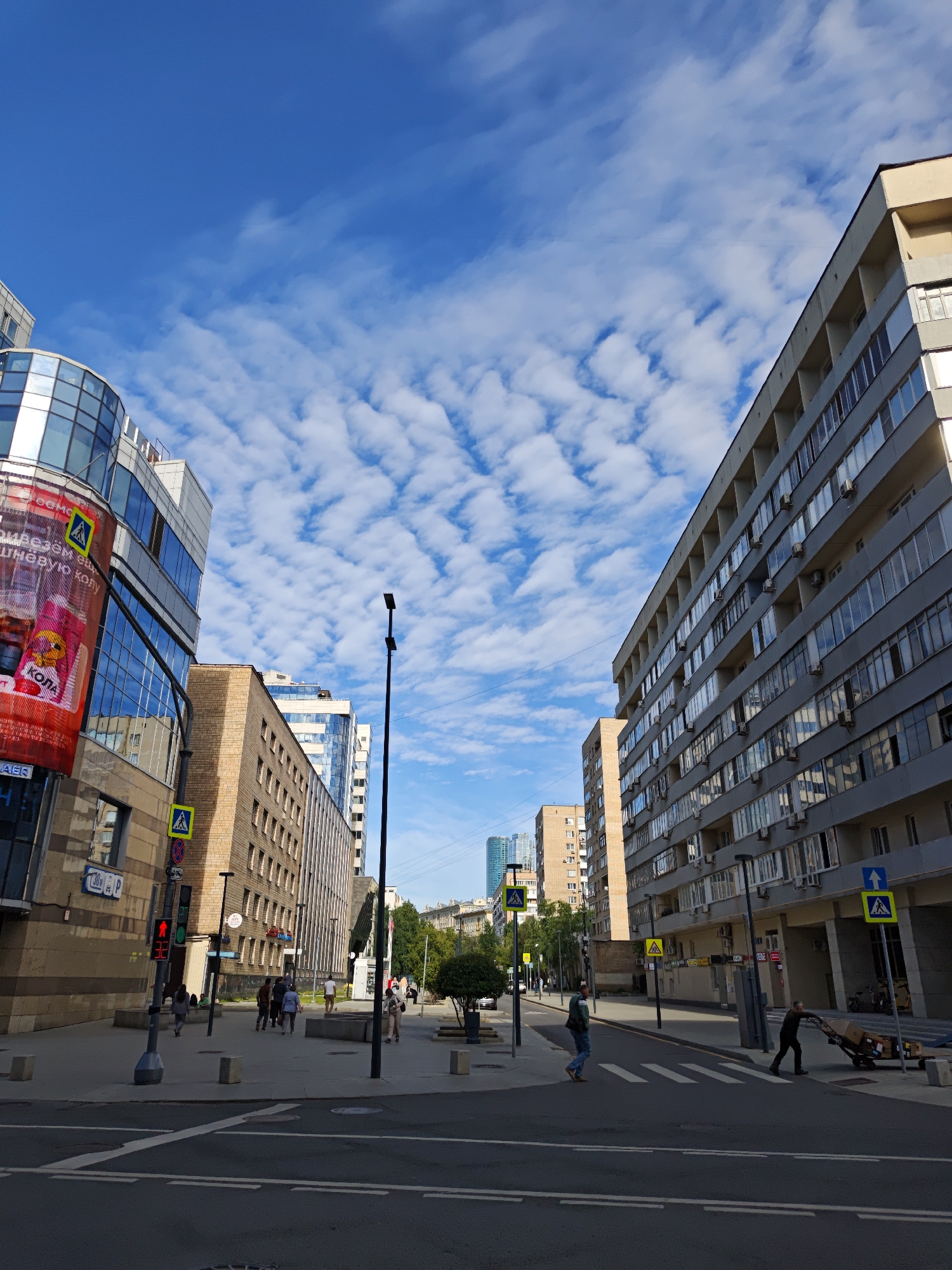 A daytime, medium-angle, eye-level shot captures an urban street scene in Moscow, Russia, beneath a bright blue sky dotted with wispy white clouds. The perspective looks down a wide street lined with modern buildings on both sides. On the left, a curved, glass-fronted building with a large red banner advertisement is prominent. Next to it, a beige brick building with multiple windows rises. On the right, a tall, grey, multi-story building dominates the view, its facade featuring numerous windows and air conditioning units.
Several pedestrians are visible on the sidewalks. Some are walking in groups, while others are crossing the street at a marked crosswalk. A man is pushing a trolley laden with cardboard boxes across the street. Traffic lights are visible, with one currently showing red. Yellow pedestrian crossing signs are posted on poles along the sidewalks. Streetlights stand at intervals. The street itself is paved, with white lines marking lanes and a crosswalk. The overall impression is of a busy, modern city environment on a clear day.