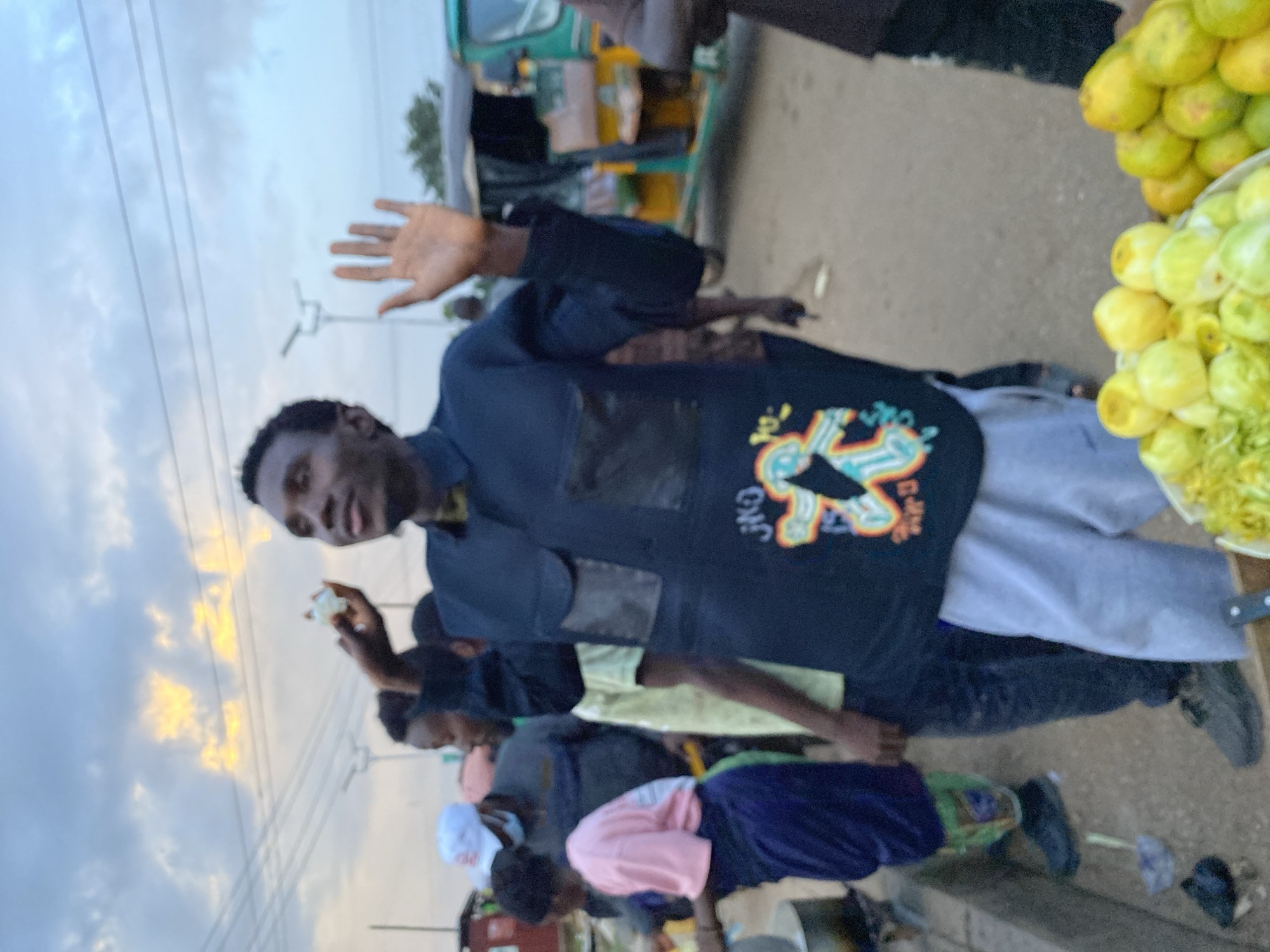 In Bukuru, Nigeria, under a cloudy sky with hints of yellow sunlight, a man stands outdoors, holding a piece of white food in his raised left hand and extending his right hand, palm open. He wears a dark long-sleeved shirt with two black rectangular patches on the chest and a colorful graphic design on the lower front. His lower body is clad in light grey sweatpants. He is positioned in a street scene with other people, a three-wheeled vehicle, and market stalls visible. To his left, a woman in a pink top and dark skirt stands by a cooking pot. Behind him, other individuals are present, and to his right, a pile of green fruits is displayed in a container, with more fruits to the right of that. The environment appears to be an outdoor market or roadside gathering.