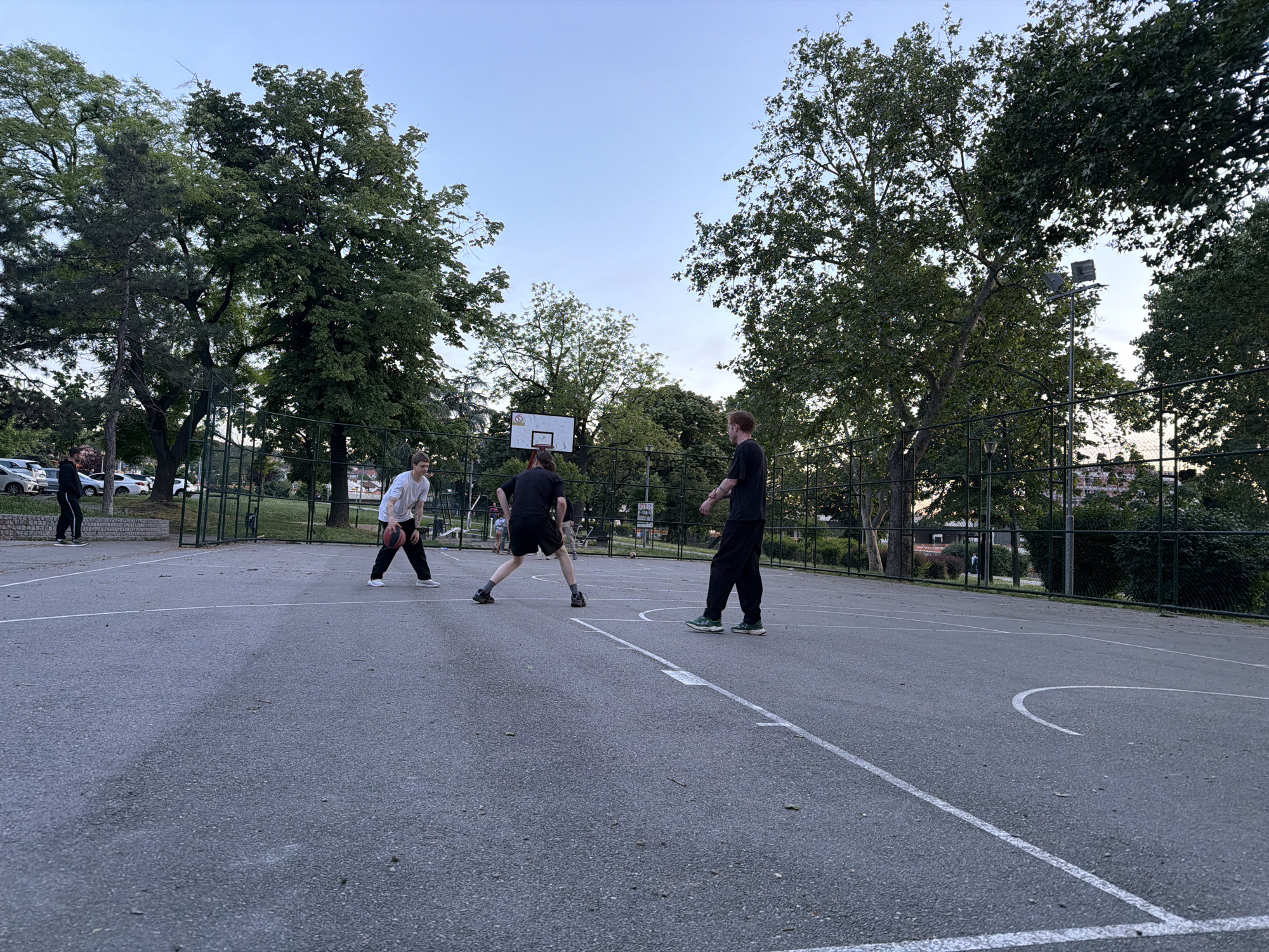 Three people are playing basketball on an outdoor court. The court has white lines painted on it. There are trees surrounding the court and a fence encloses the area. The sky is a light blue color. In the background, there are parked cars, and buildings can be seen in the distance. There's a streetlight visible near the treeline. The photograph seems to be taken at Tašmajdan Park in Belgrade, Serbia.