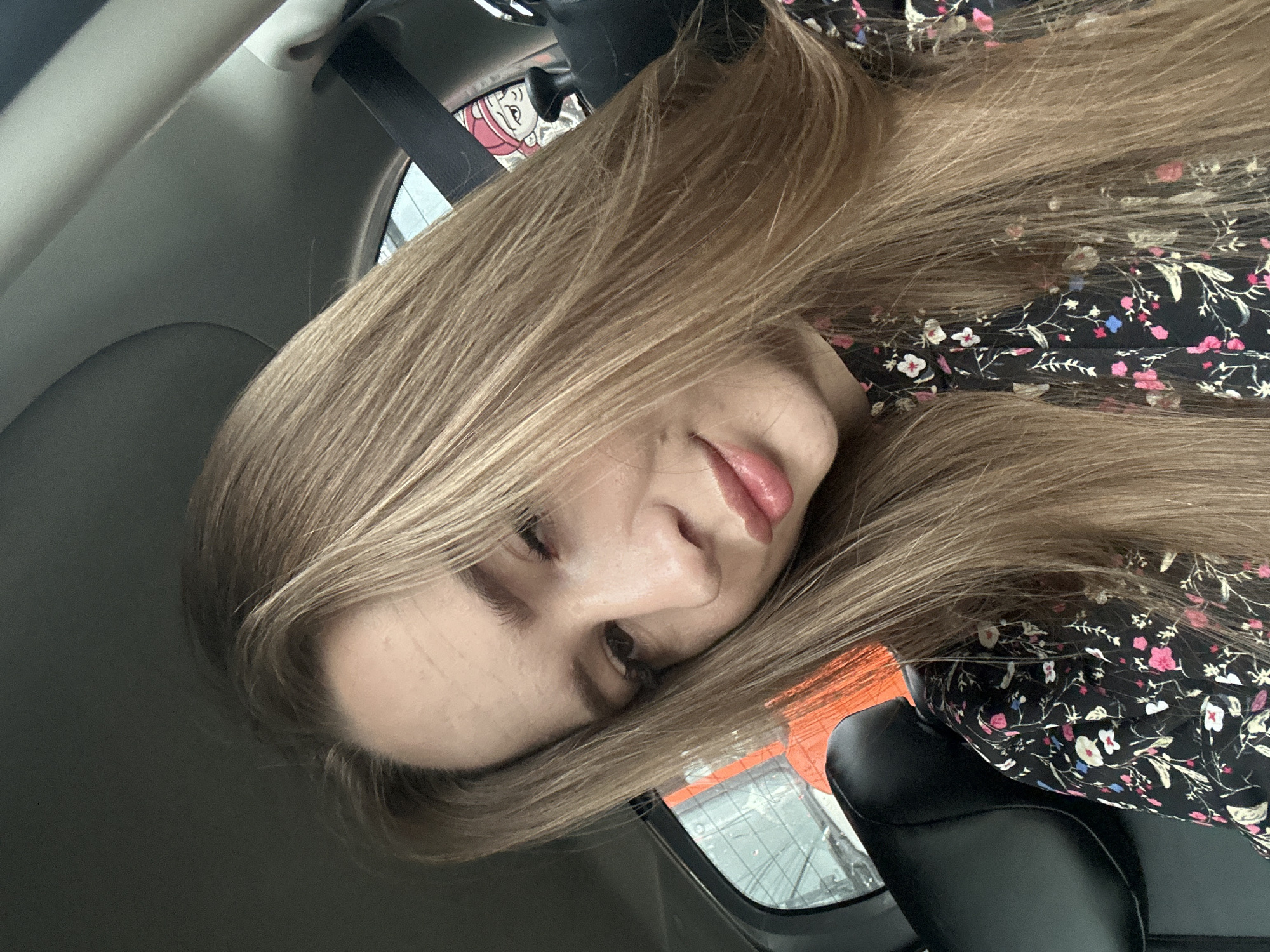 A woman with long, blonde hair is sitting in the passenger seat of a car. She is wearing a black dress with a floral pattern. The woman is looking at the camera and smiling slightly. The car appears to be moving, as indicated by the blurred background. The overall lighting suggests it is daytime.
