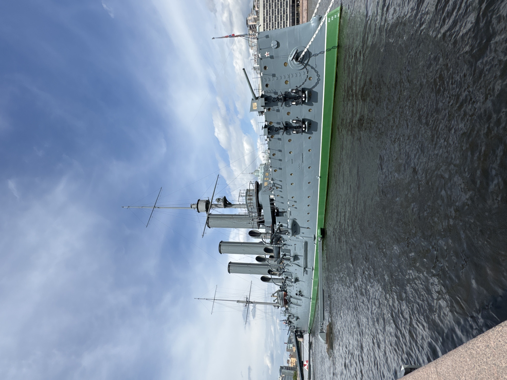 The image displays the port side of a large, grey naval cruiser, likely the historic Aurora, permanently moored on a body of water in Saint Petersburg, Russia. The ship occupies the central and left portions of the frame, with dark, rippled water filling the lower part and a blue sky with scattered white clouds above.
The cruiser features a grey hull with a distinctive bright green waterline stripe, edged by a narrow white band. Its deck is equipped with multiple large gun turrets, several smaller anti-aircraft guns, and a complex array of masts, rigging, and three prominent smokestacks. An anchor and heavy chain are visible at the bow. A red star emblem is clearly depicted on the forward section of the hull, and a flag, possibly the Russian naval ensign, flies from a staff near the bow. On the green waterline stripe, white Roman numerals "XXX AX" and "XIV XXXI" are visible towards the front of the ship.
The dark water reflects the sky and portions of the ship. Across the water, on the far bank, several buildings of the Saint Petersburg skyline are visible, including a notable multi-story structure. The immediate foreground on the right side of the image shows the textured surface of a granite or stone embankment. The bright blue sky and fair-weather clouds indicate a clear day, likely late morning or afternoon. No people are visible on the ship or quay. The scene evokes a sense of historical significance and peaceful grandeur, characteristic of a museum ship display.