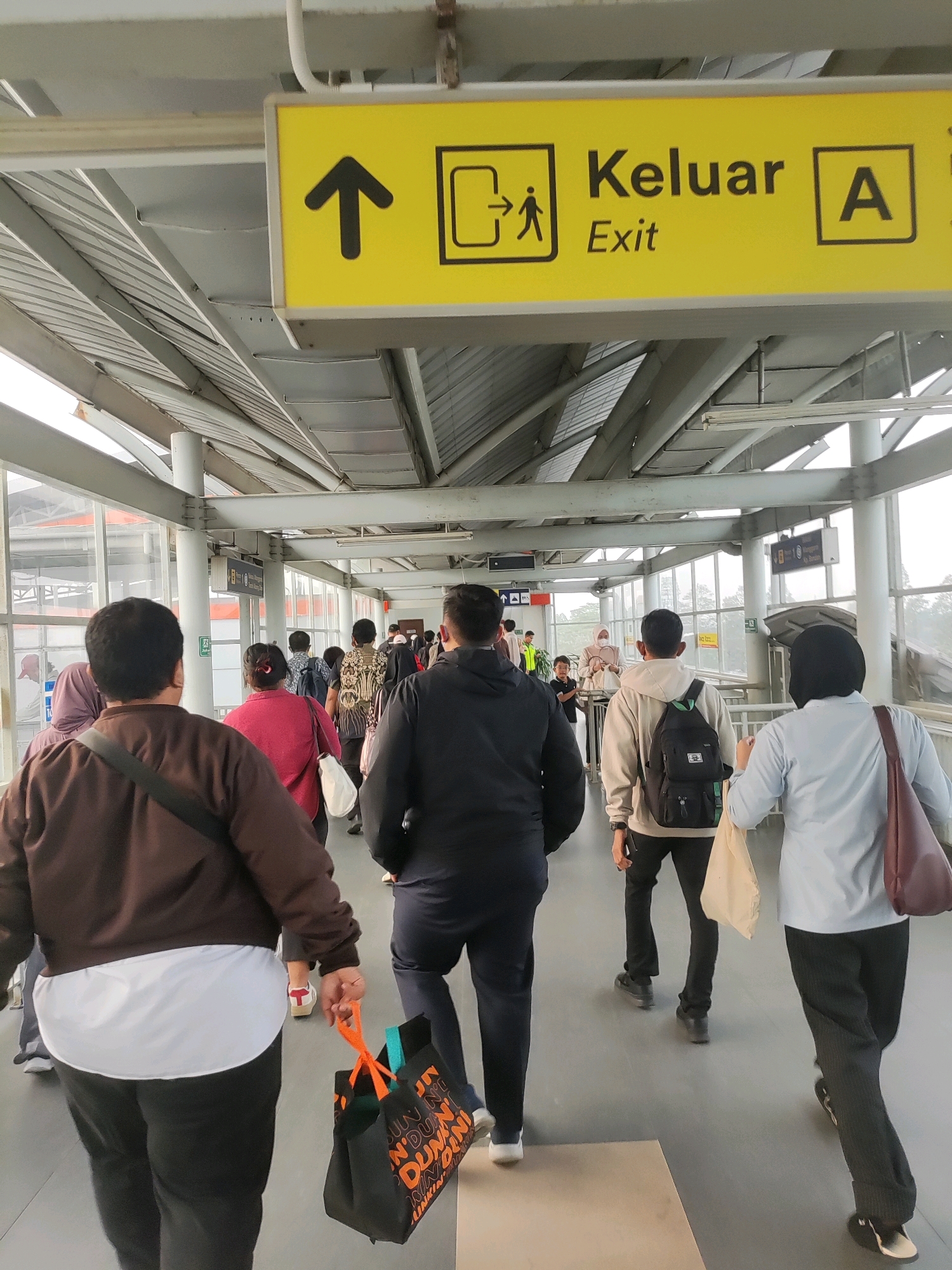 In Telagamurni, Indonesia, a group of people are walking along an elevated walkway, likely at a train station or transportation hub. The walkway is covered by a corrugated metal roof supported by metal beams, and has glass panels on the sides. A prominent yellow sign with black lettering hangs overhead, indicating "Keluar Exit A" with an icon of a person exiting a doorway. People are dressed in casual attire, with some carrying bags. The scene depicts a typical flow of commuters.