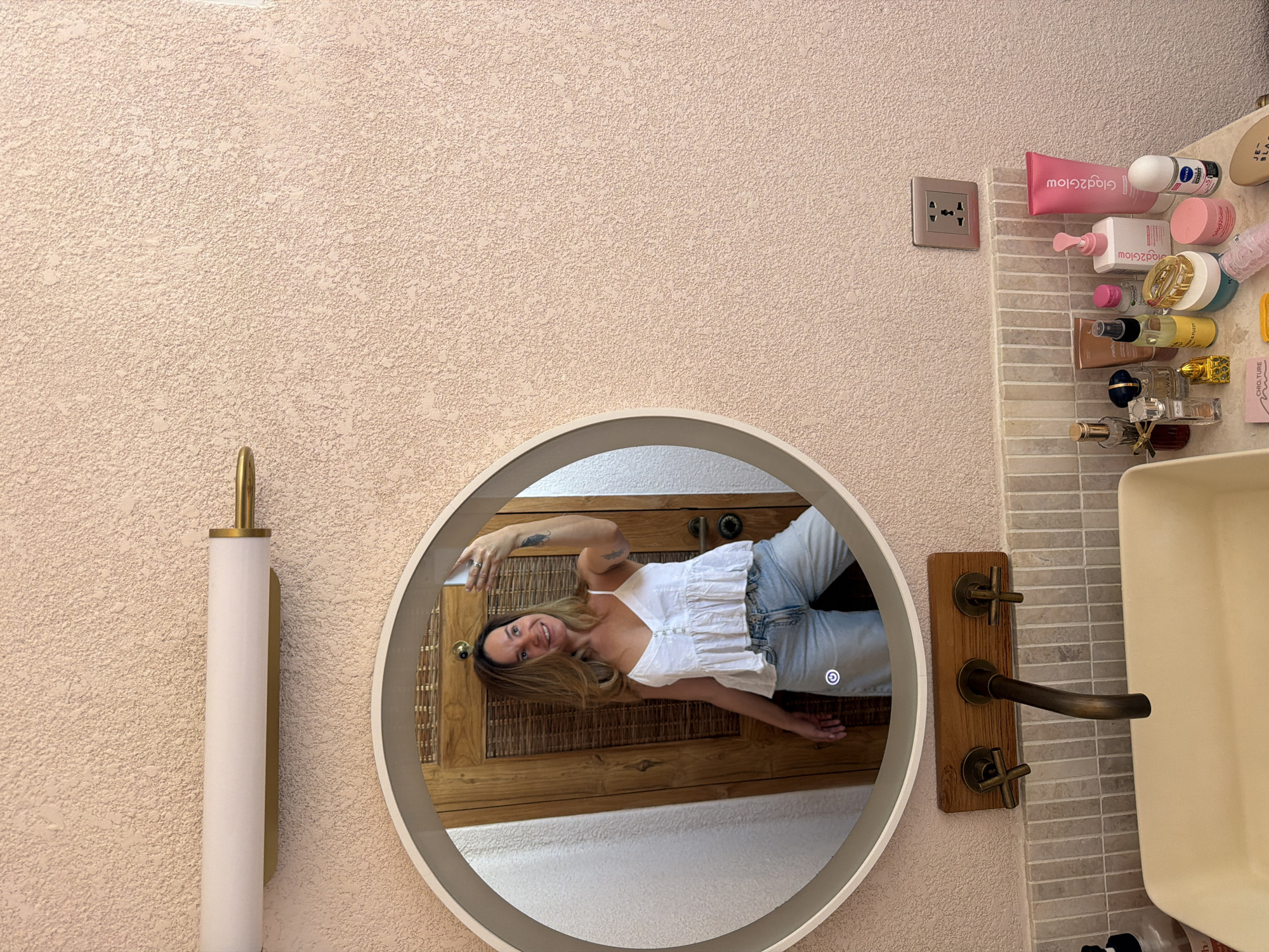 This media file captures a woman taking a selfie in a bathroom, reflected in a circular, illuminated mirror. The woman has long brown hair, is smiling, and wears a white sleeveless top and light blue jeans. She holds a smartphone to capture her reflection.
The setting appears to be a modern yet rustic bathroom in Mengwi, Indonesia. The wall has a light beige, textured finish. To the left of the mirror is a contemporary wall sconce with a gold base and a long, white cylindrical shade.
To the right of the mirror, a vanity area features a light-colored, rectangular sink. Above the sink, a unique brass or bronze faucet assembly with two cross-handle valves is mounted on a wooden plate. The wall behind the sink is tiled with small, light-colored rectangular tiles. Numerous toiletries are arranged on the countertop, including tubes, bottles, and jars. Visible text on some products includes "Giga2glow" (reversed), "Nivea", "Chio ture", and "JF". A silver multi-socket power outlet is visible on the textured wall above the toiletries.
The background reflected in the mirror behind the woman shows a wooden door with a woven panel and what appears to be a light-colored floor or rug. The woman's joyful expression, combined with the personal items, suggests a comfortable and private setting.