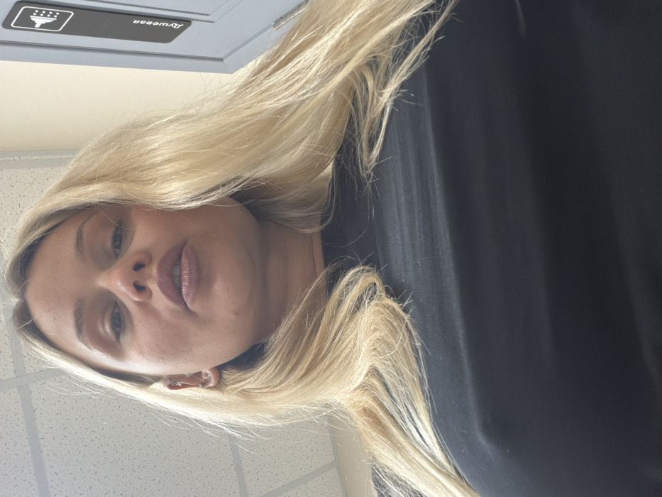 A low-angle indoor image depicts a blonde female subject looking directly into the camera. She has long, straight blonde hair, multiple piercings in her left ear, and is wearing a black top, through which the outline of her left nipple is visible. Her lips are slightly parted. The background features a white drop-tile ceiling in the upper left. To the upper right, a light-colored wall displays a vertical, grey rectangular sign with a white shower icon at the top and the Cyrillic word "Душевая" (Dushevaya), translating to "Shower room," written vertically below. The scene is located in a facility within the city of Sarapul, Russia.