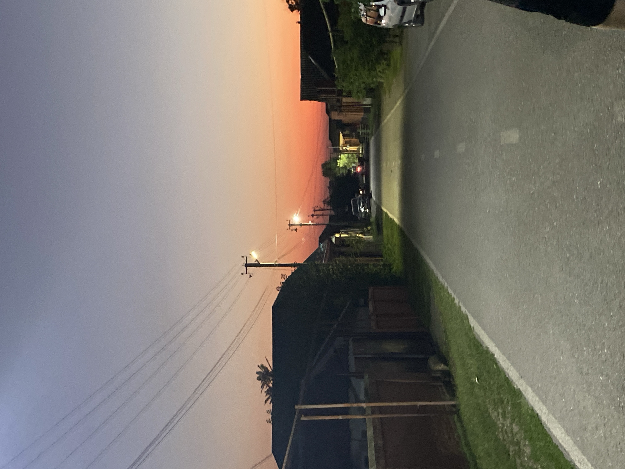 The image depicts a street scene in Alkhan-Kala, Russia, during twilight. The sky transitions from a pale blue at the top to a vibrant orange and pink hue near the horizon. Power lines stretch across the upper portion of the frame. On the left side of the street, a dark-colored building with a sloped roof is visible, bordered by a brick wall and grass. Further down the street, more buildings line both sides, some with visible roofing and windows. Streetlights are illuminated, casting a glow on the asphalt road. Several cars are parked or driving on the street, including a silver SUV on the right side. The overall impression is that of a quiet, residential street at the end of the day.