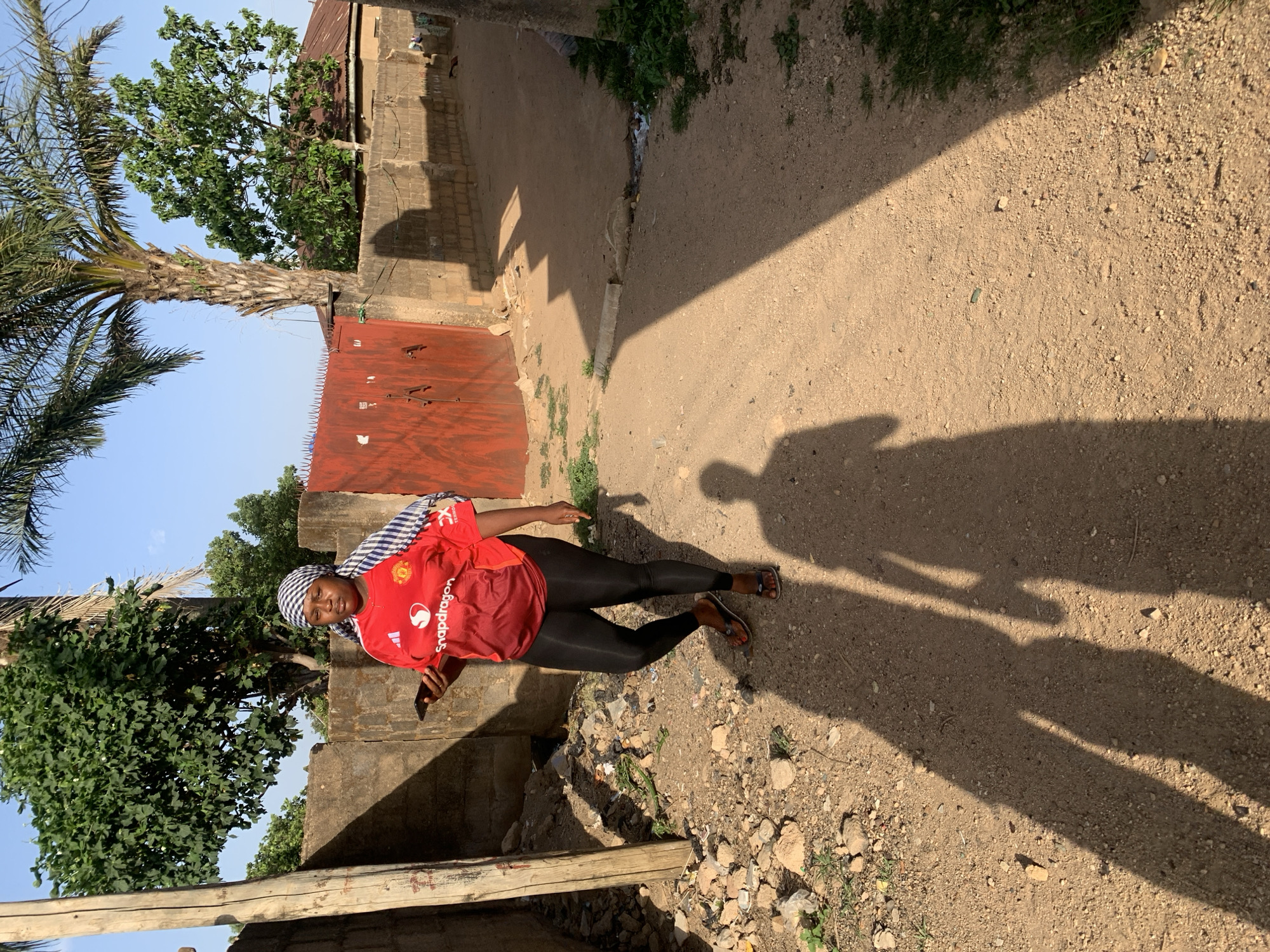 A woman stands on a dirt path in Jos, Nigeria. She wears a red Manchester United jersey, black leggings, and sandals. Her head is wrapped in a patterned scarf. In her hand, she holds a dark-colored mobile phone. The path is lined with a crumbling concrete wall on one side and a red corrugated metal fence on the other. The shadow of the woman and another individual is cast on the dirt path. Trees and buildings are visible in the background.