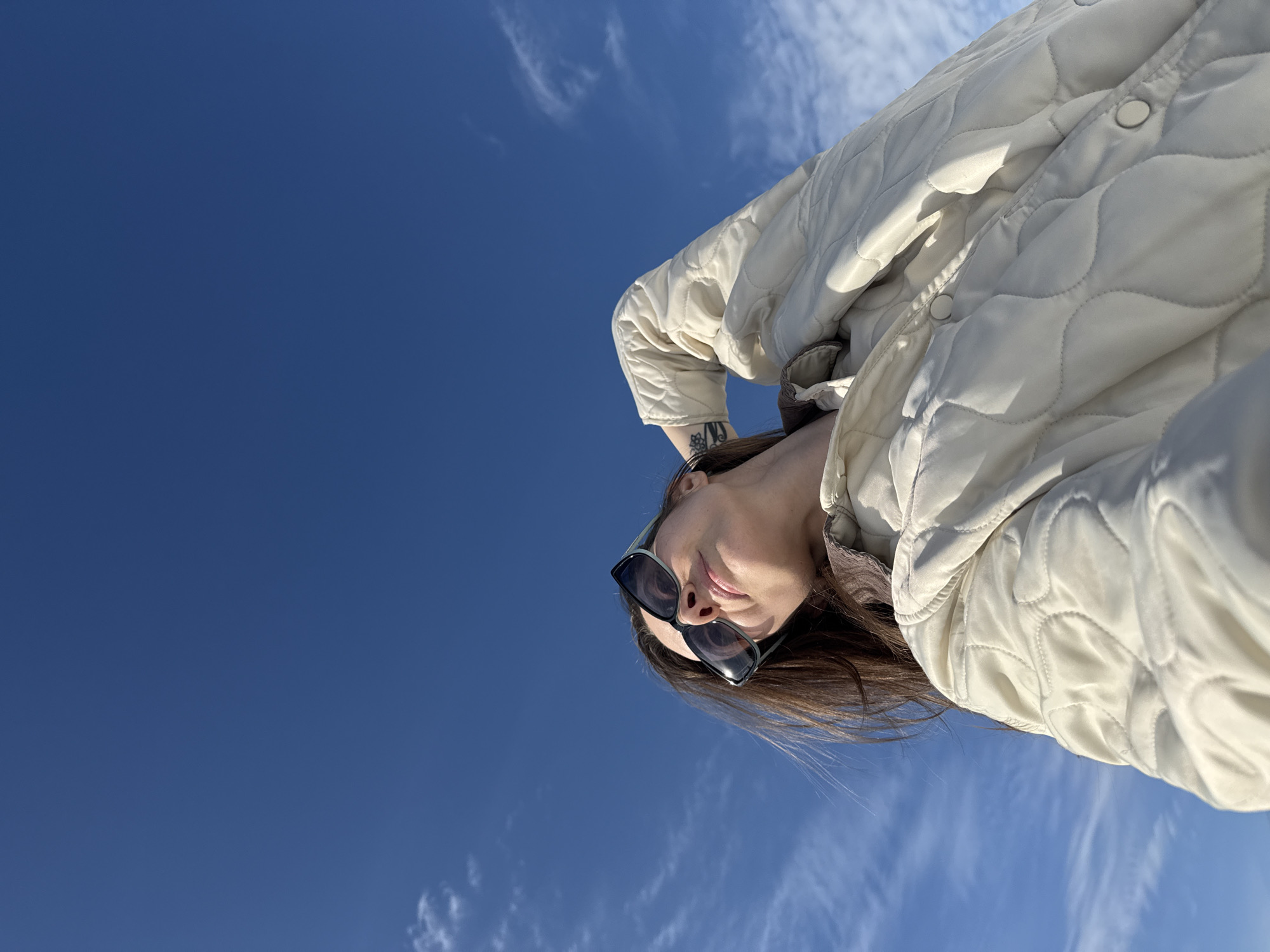 A low-angle photograph captures a woman gazing upwards against a clear blue sky, featuring scattered wispy cirrus clouds. She wears dark-framed sunglasses and an off-white, quilted jacket with visible snap buttons, and a brown collar underneath. Her dark hair is parted and slightly windblown. Her right arm is bent and raised, with her hand positioned behind her head, revealing a black tattoo on her right forearm. The scene is situated in the city of Chaikovsky, Russia.