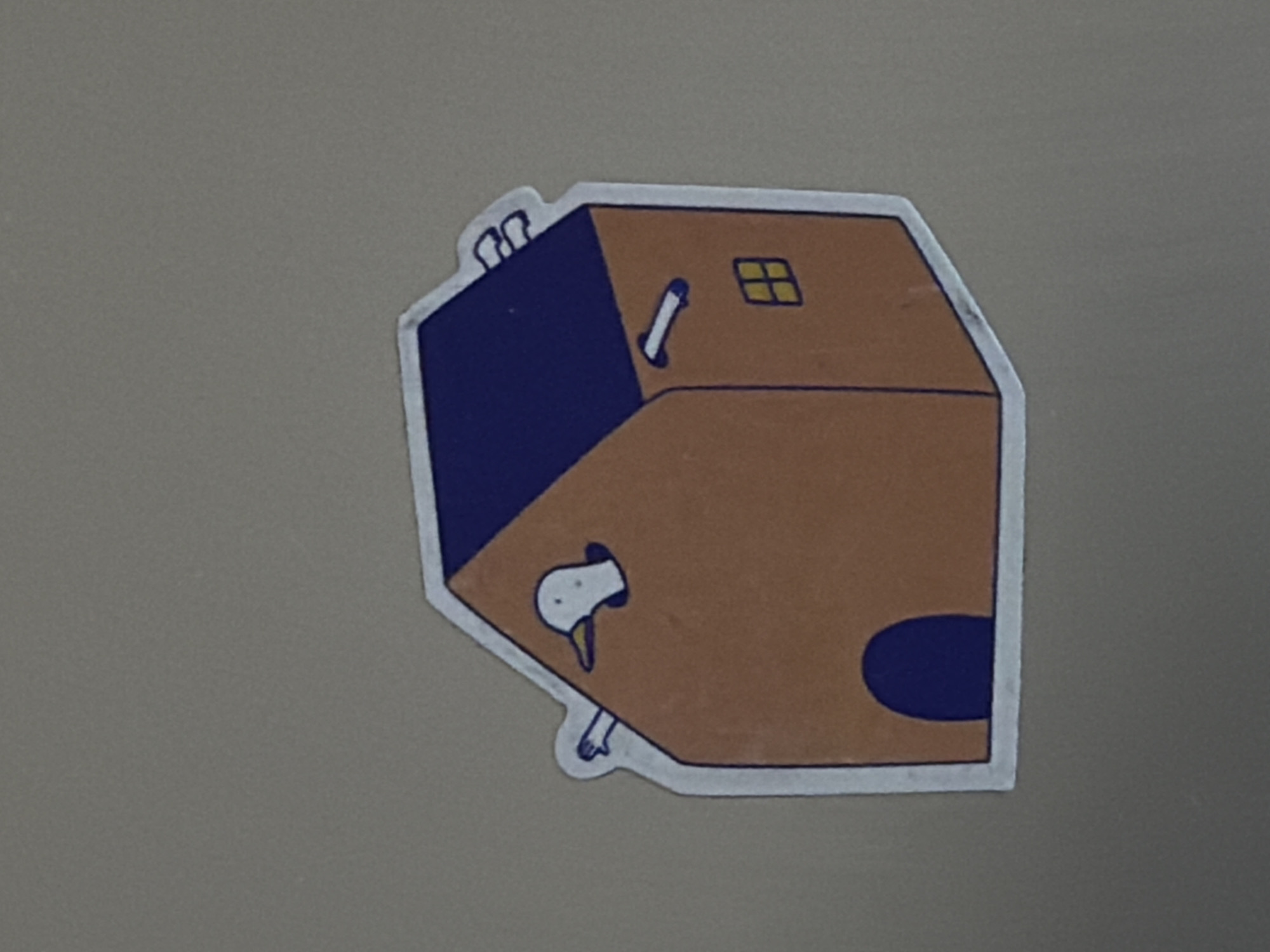 The media file contains a sticker with a cartoon illustration. The main subject of the sticker is a stylized brown box with a blue top. On the front of the brown section of the box, there is a drawing of a white bird's head peeking out of a hole. The bird has small black eyes and a yellow beak. Two white bird legs are also visible, one sticking out from under the box and another within the hole. On the top blue section of the box, there are two white cylindrical shapes that resemble chimneys. A small yellow square with black lines, representing a window, is visible on the top brown side of the box. A dark blue oval shape is present on the right side of the brown section, potentially representing a door or another opening. The sticker has a white border around the illustration. The background of the media file is a plain, neutral-toned surface, likely a wall or a table. There is no discernible text in the image. The overall style is simple and whimsical, suggesting a lighthearted or playful theme.