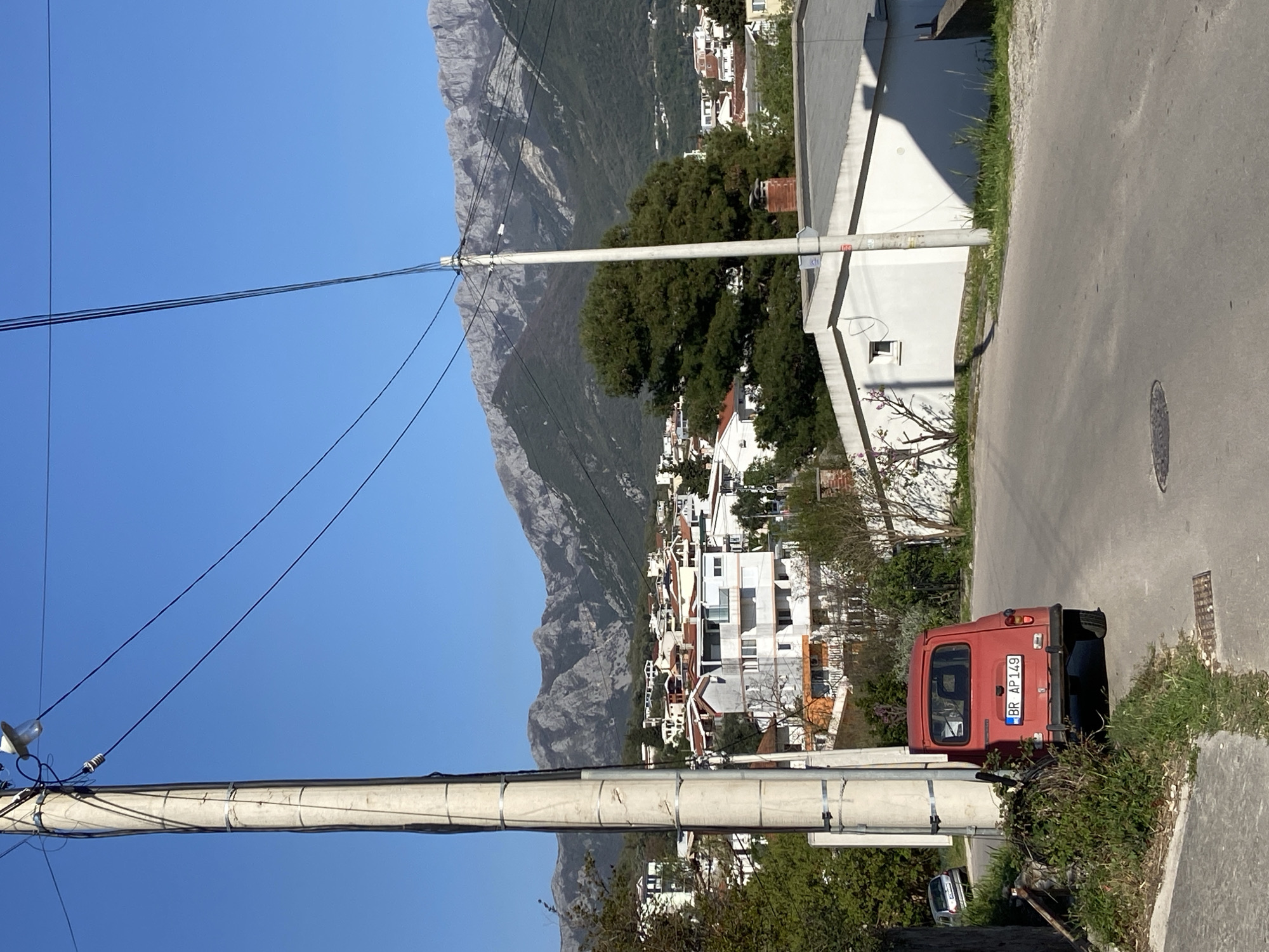 The image captures a street scene in Šušanj, Montenegro. The focus is on a parked, vintage, red car with license plate "BR AP 149", positioned on a paved road partially overgrown with vegetation. To the left of the vehicle are utility poles with power lines cutting across the clear blue sky. In the background, a hillside densely populated with white buildings and residences ascends toward a rugged mountain range. A white building runs alongside the road, parallel with the car.