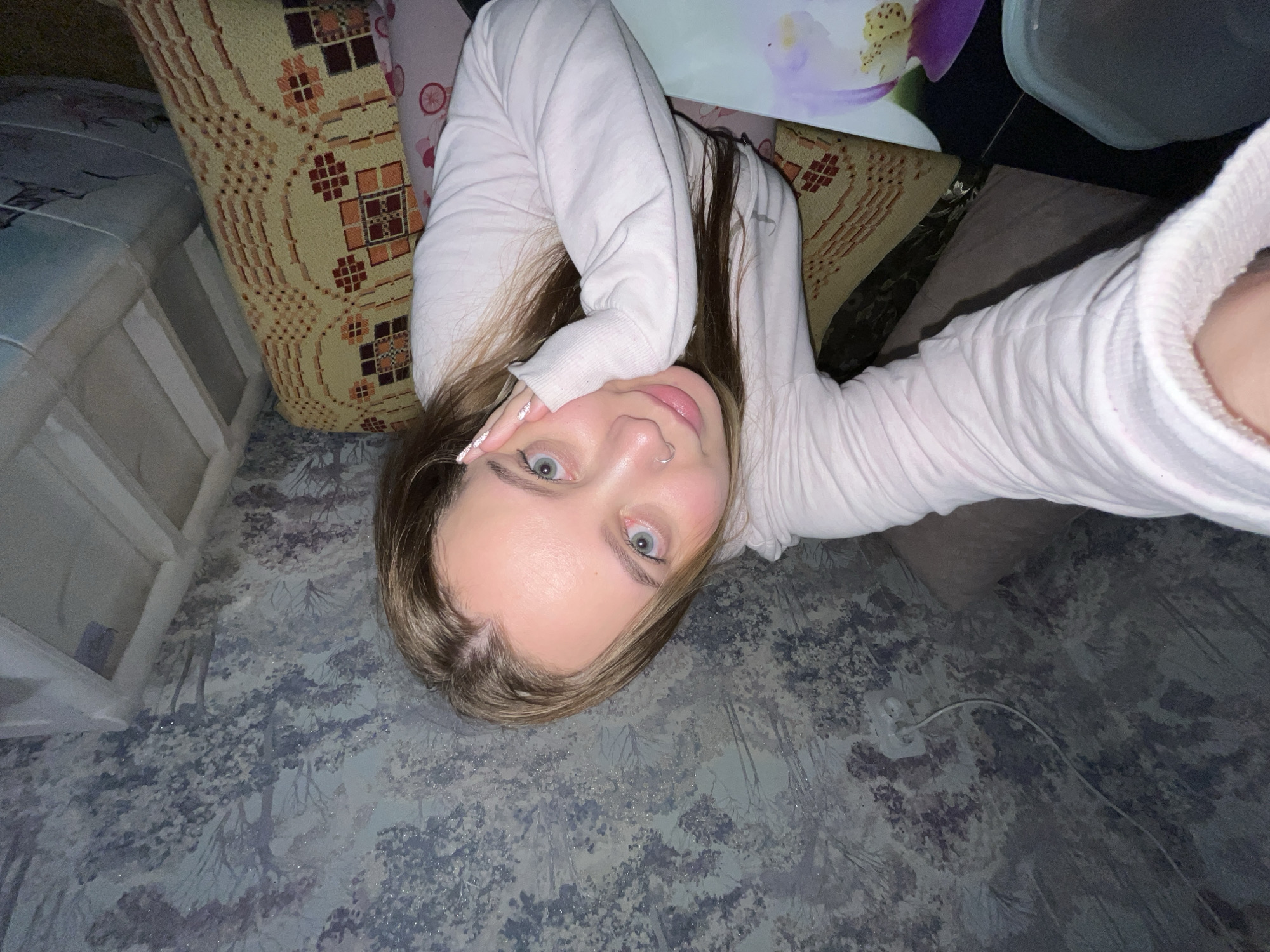 A young woman lies on a patterned floor in what appears to be an indoor setting, likely a room in a residence. She is positioned in the center of the frame, gazing directly at the camera with light blue eyes. The woman wears a light-colored, long-sleeved garment. Her hair is a mix of brown and blonde hues, styled long and loose. Her left hand is raised and resting on her forehead. The background features a variety of textured surfaces, including pillows with geometric patterns, furniture, and walls covered in a blue and gray nature-inspired pattern. An electrical outlet with a cord is visible near the bottom right corner of the image. The overall lighting is dim. This image was taken in Novopokrovskaya, Russia.