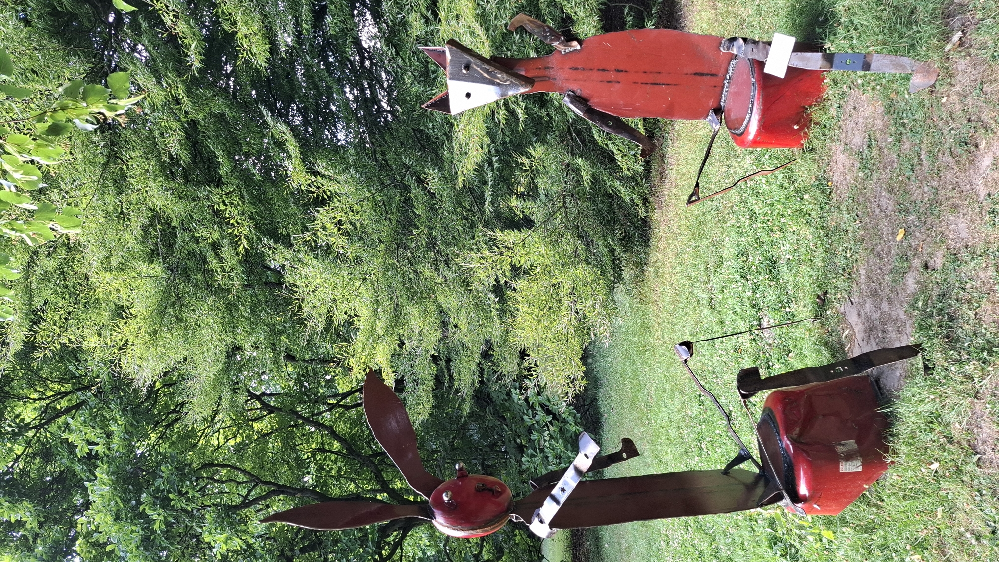Two metal sculptures of stylized animals are situated on a grassy area with trees in the background. The sculpture on the left appears to be a rabbit, made of dark red metal with a red, round head that has silver accents for eyes. It is sitting on a red, rounded object, possibly a repurposed drum or barrel, and its arms are bent at the elbows, extended forward. The sculpture on the right is a fox-like creature with a triangular head that is black and white, and a red body. It is also sitting on a similar red, rounded object, and its arms are bent and extended forward. Both sculptures are constructed from metal, with visible seams and welded joints. The ground around the sculptures is covered in green grass, with patches of dirt. The background consists of lush green trees, indicating an outdoor, natural setting. The overall scene suggests an artistic installation in a park or garden environment. This image is located in the sculpture park of the German Sport University in Cologne, Germany.