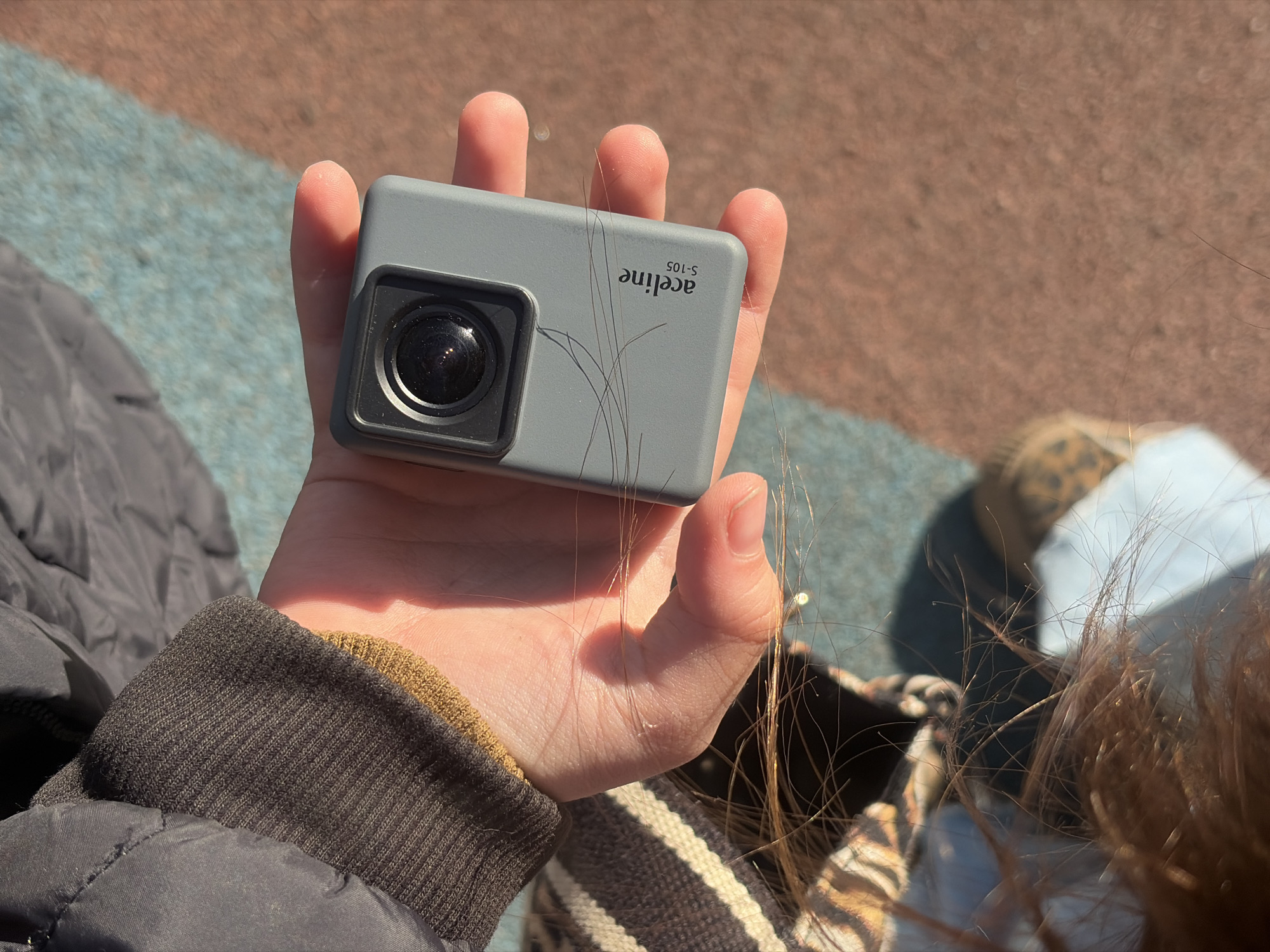 A hand holds a gray action camera labeled "aceline S-105." Strands of brown hair are intertwined with the fingers holding the camera. The person holding the camera wears a black jacket with a ribbed brown cuff. Partially visible in the background are a striped bag, a shoe with a leopard print, a light blue surface, and brown ground. The scene is lit by natural sunlight. The location is Saint Petersburg, Russia.