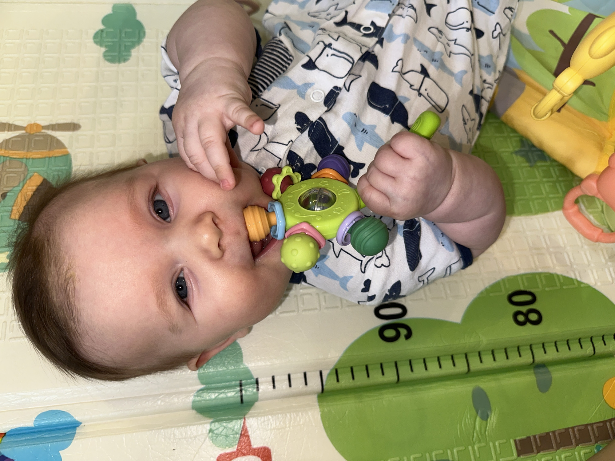 A baby lies on a patterned play mat. The baby wears a white shirt with shark patterns. The baby holds a green, yellow, and purple toy. The mat has images of clouds, an airplane, and a ruler marked in centimeters. The numbers 80 and 90 are visible on the ruler. The location and city are not evident in the image.