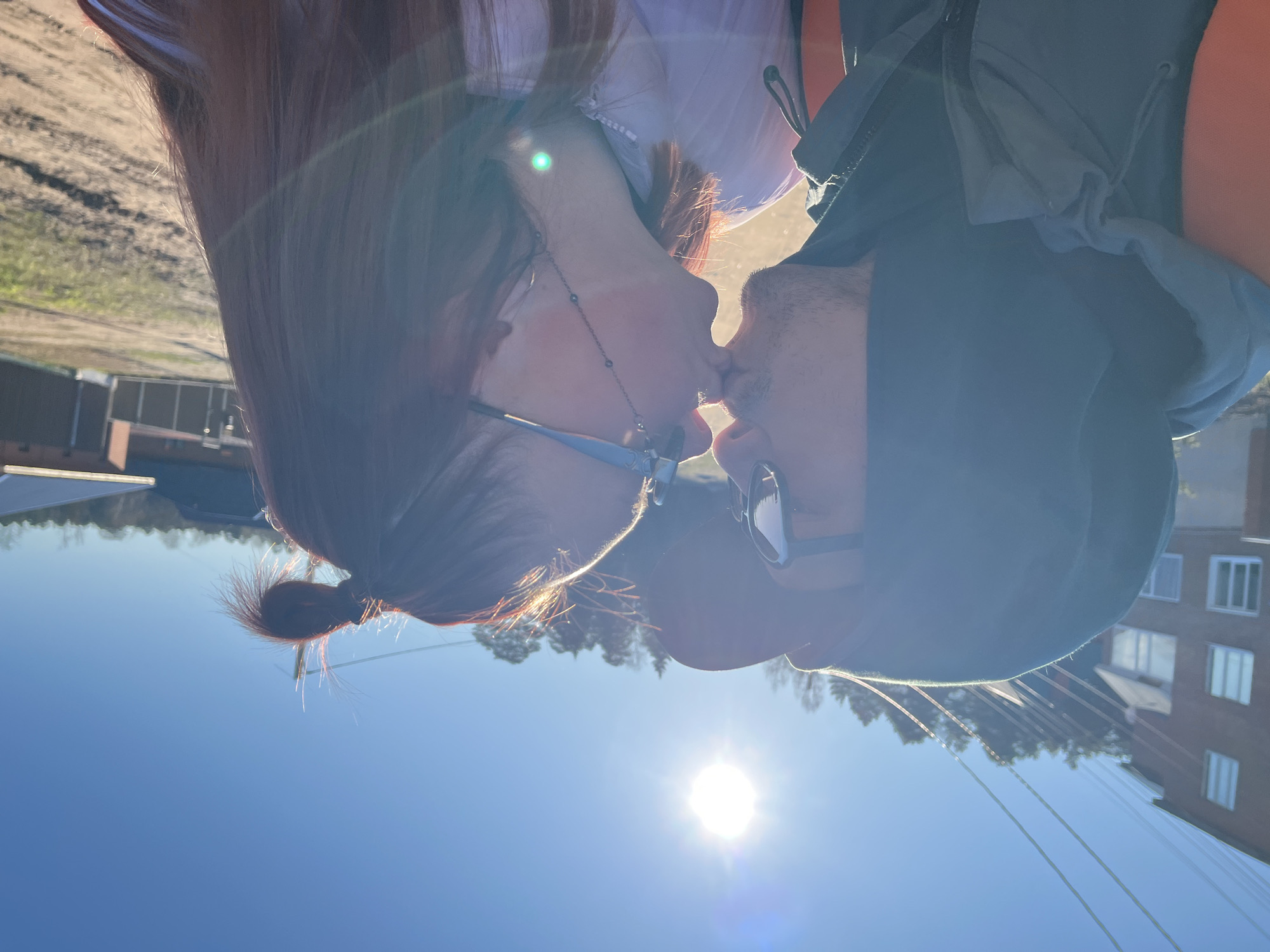 The image shows two people, likely a couple, captured in a tender moment as they kiss outdoors in Chaikovsky, Russia. The photograph is taken from a low angle, looking upwards, with the bright sun positioned towards the lower right, casting a strong flare and backlighting the subjects. The sky is clear and blue, indicating sunny weather, probably during midday or early afternoon.
On the left side of the frame, a woman with long, reddish-brown hair is seen. Her hair is partially pulled back into a bun, visible towards the bottom of her head. She wears blue-framed prescription glasses and a light-colored top, possibly white or light blue. A delicate beaded necklace with a small pendant hangs around her neck.
On the right, a man wearing a dark, possibly grey or green, hooded jacket and sunglasses leans in to kiss the woman. Their lips are touching, emphasizing the intimate nature of the interaction. The strong sunlight illuminates the outlines of their profiles and hair, creating a warm glow.
The background features a mix of urban and natural elements. In the upper left, a patch of what appears to be a dirt road or pathway with some greenery is visible. Further down and across the background, silhouettes of trees are seen against the sky. On the far right, a multi-story building, possibly an apartment block with several windows, is discernible. Overhead power lines also crisscross the sky on the right side of the image. The scene conveys a sense of affection and connection between the individuals amidst the bright outdoor setting. There is no visible text in the image.