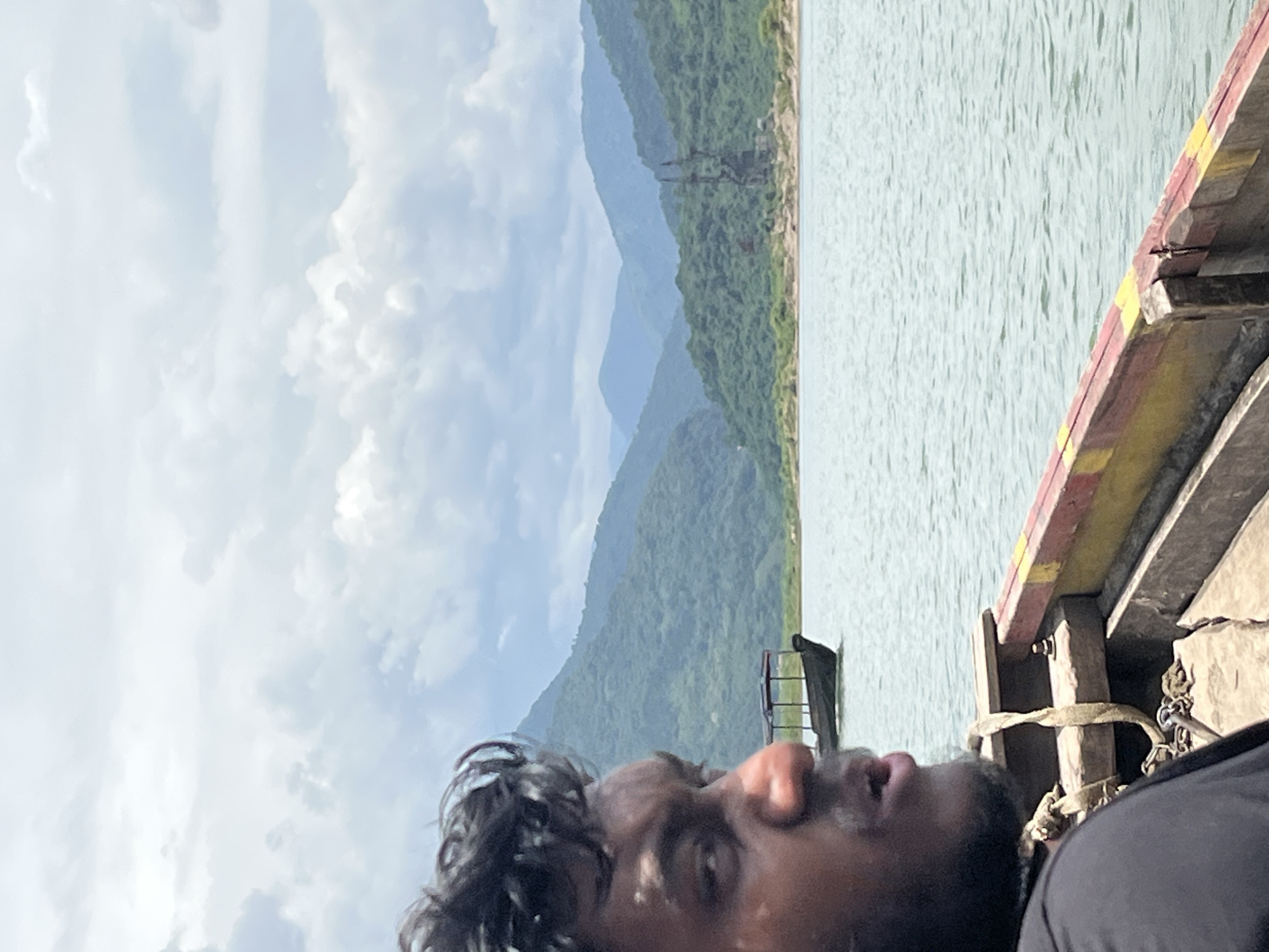 The image depicts a young man in the foreground, facing left and looking off into the distance. He has dark, curly hair and is wearing a black t-shirt. He appears to be on a boat, as indicated by the wooden railing with red and yellow paint visible in the lower right portion of the frame. In the background, a body of water stretches out towards a lush, green mountain range under a cloudy sky. Another boat with a canopy is visible on the water in the middle ground, and in the far distance, a tall, metal structure, possibly a pylon or transmission tower, stands against the backdrop of the mountains. The location is identified as Majai, India.