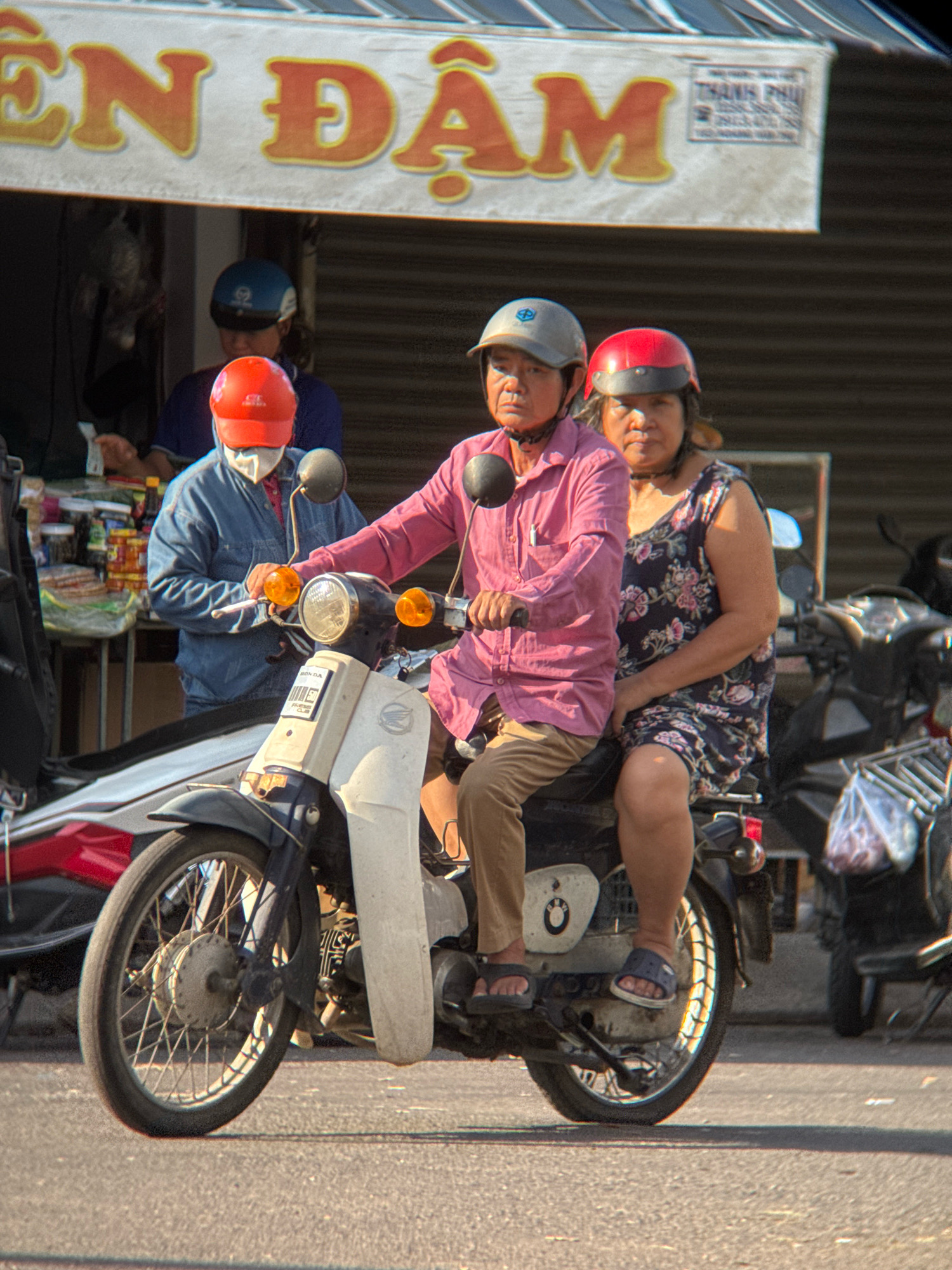 A medium shot captures two individuals on a motorcycle in Nha Trang, Vietnam. The man in front wears a pink collared shirt and tan pants, with a grey helmet and a white mask partially obscuring his face. He holds the handlebars, his left hand on the throttle. The woman behind him wears a dark floral print dress and a dark helmet. She is seated, her hands resting on her lap. The motorcycle is white with blue accents. In the background, a store with a striped awning displays signage with Vietnamese characters, and shelves stocked with various items are visible. The setting appears to be a street or thoroughfare, with a paved surface visible in the foreground.
