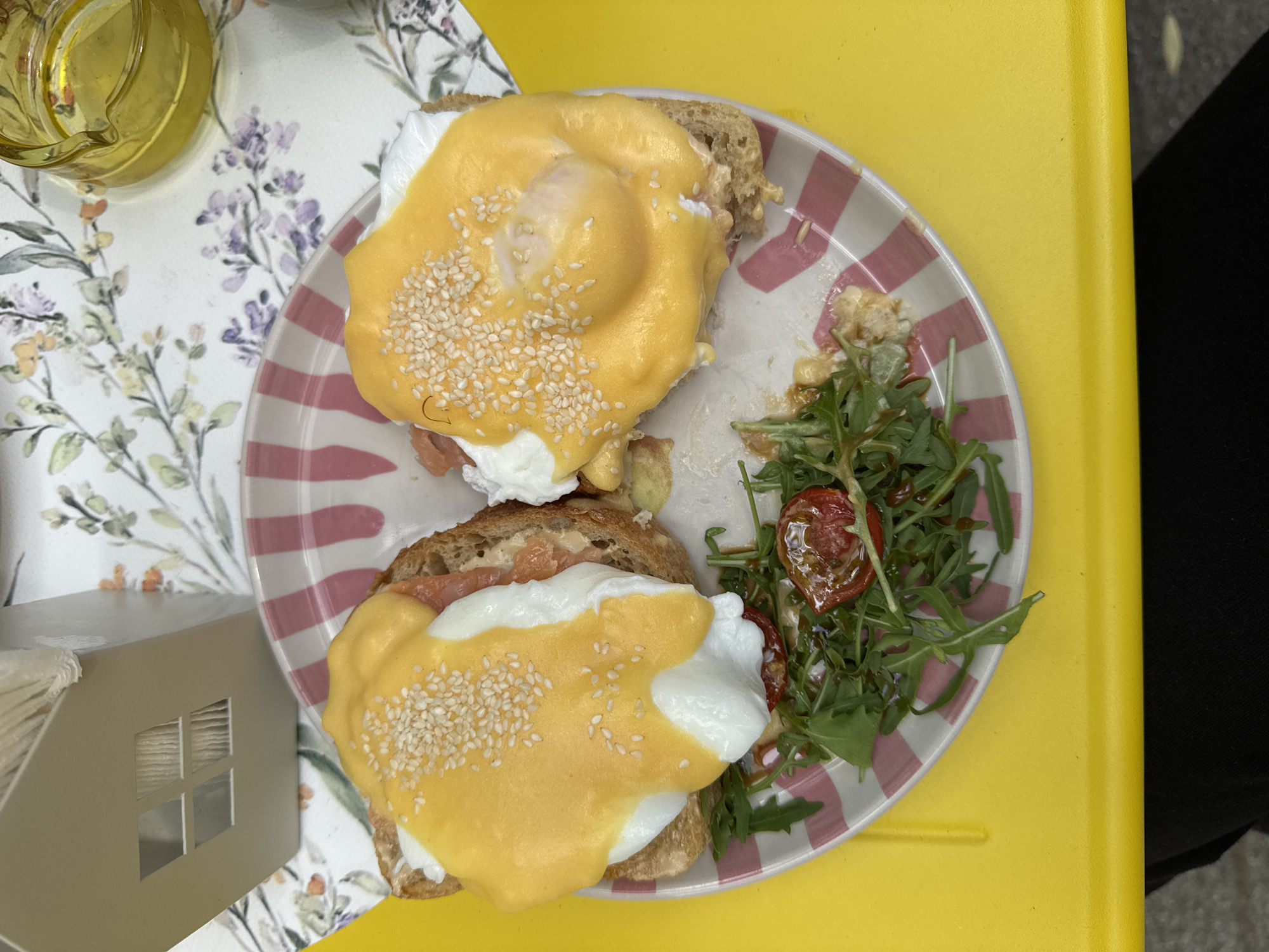 A top-down shot shows a striped plate with two open-faced sandwiches topped with a yellow sauce, white egg, and sesame seeds, along with a side of arugula and tomatoes. The plate is set on a yellow surface to the right, while a floral-patterned surface is visible on the left, with a yellow glass object and a tissue holder. A person wearing black pants is to the right of the plate. The location in the city is Unknown, Unknown.