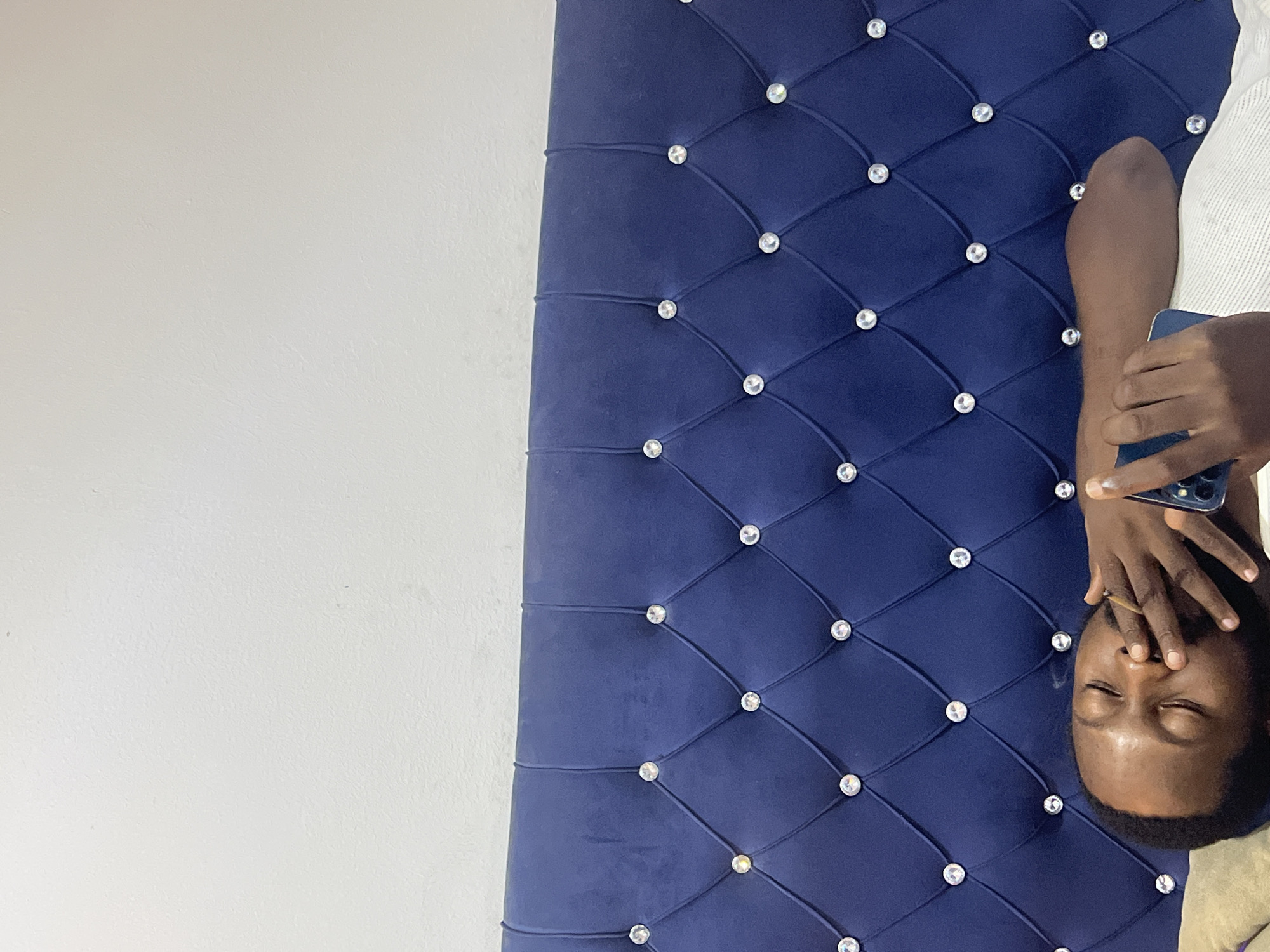 A man lies on a blue, jeweled headboard in front of a white wall. He is holding a phone and his hand is near his face. It is not possible to identify the specific location within Abuja, Nigeria from the image.