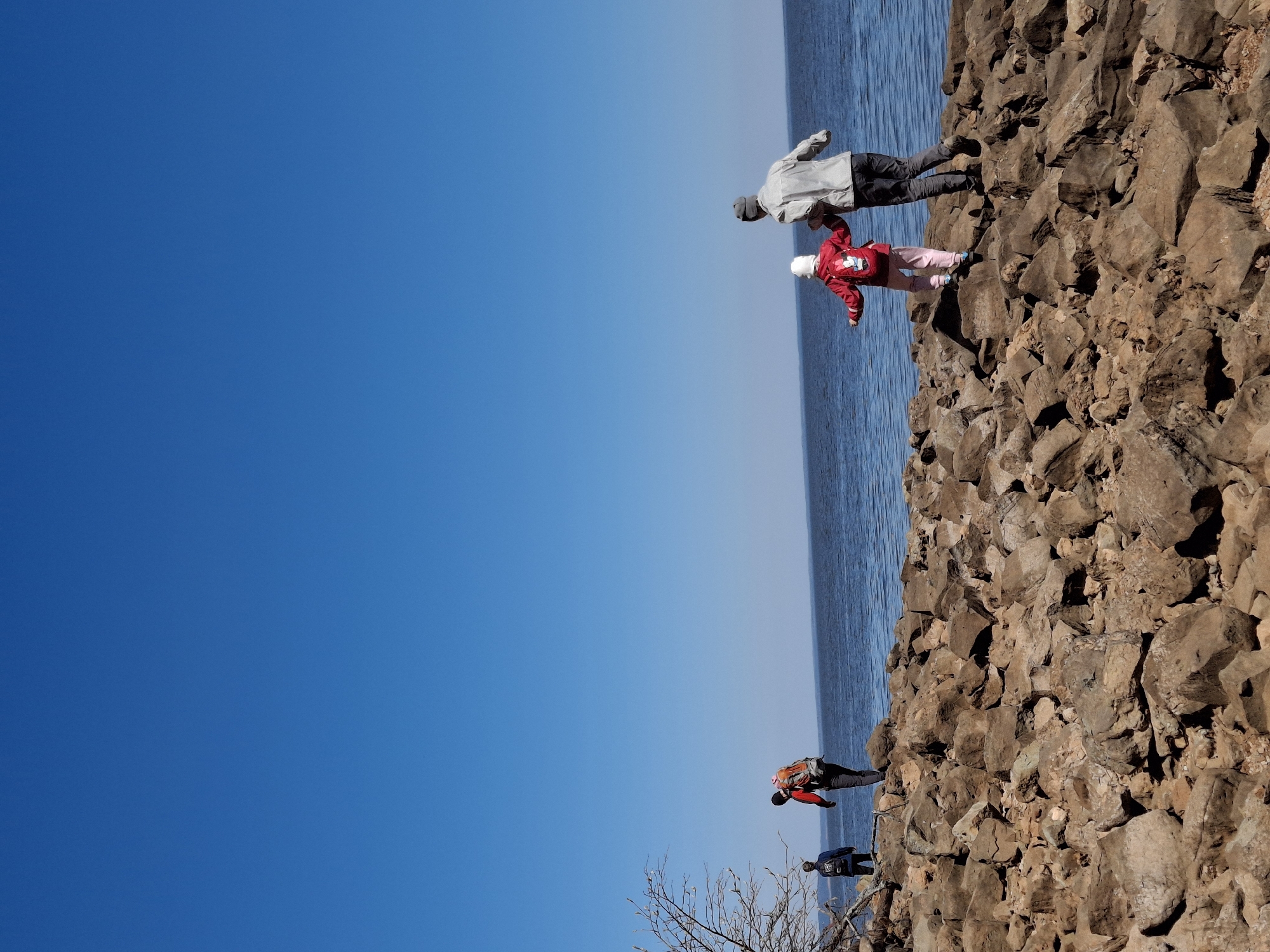 A rotated medium-angle shot shows several people traversing a rocky terrain, likely a coastal area, under a clear, blue sky.
In the upper part of the frame, two people are visible: an adult in a light-colored jacket and dark pants is helping a child in a red jacket and pink pants walk across the rocks.
Further down the frame, another person with a backpack is on the rocks. At the bottom of the frame, a man in black clothing with a backpack is walking towards the bare branches of a tree.
The rocky terrain transitions into the ocean, which has a slight blue-grey hue. The sky occupies a significant portion of the frame and is a solid, bright blue.