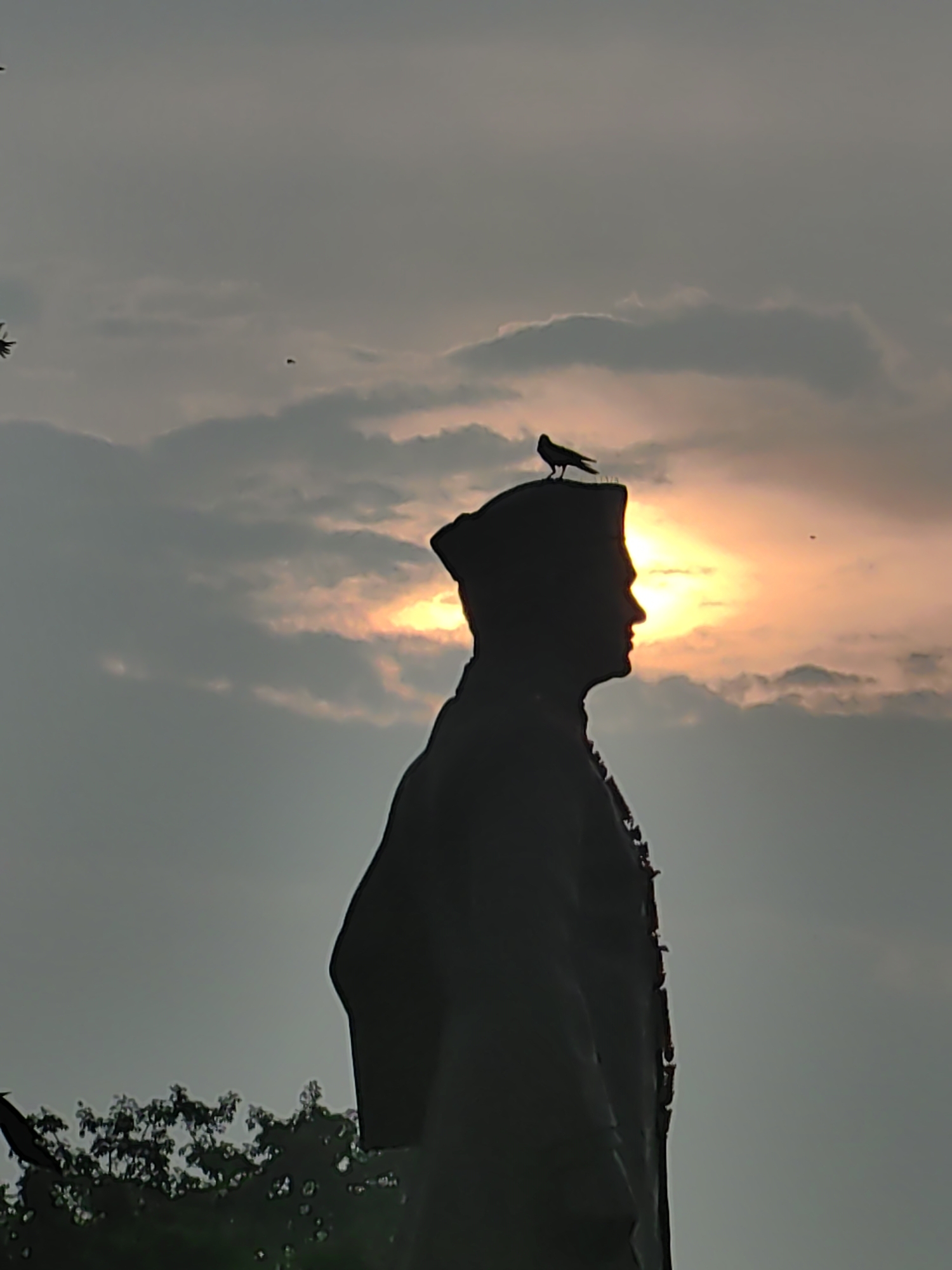 A silhouette of a statue stands against a cloudy, orange-hued sunset sky. A bird is perched on the head of the statue. The statue is of a man wearing a cap and a jacket. The statue is located in the city of Pura Raghunathpur, India. The sun is partially obscured by clouds, casting a warm glow. The sky is a mix of gray and orange, with some darker clouds present. In the foreground, the silhouette of trees can be seen at the bottom of the image.