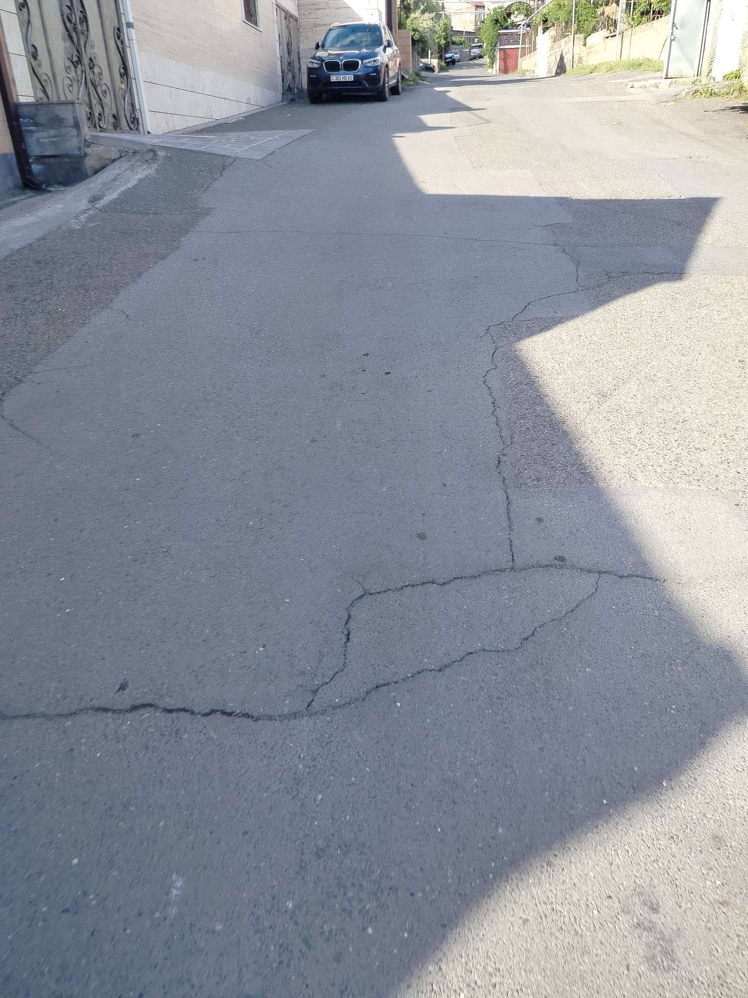 The image shows an asphalt street in Yerevan, Armenia. The road surface has several cracks. A dark blue BMW SUV with license plate "301 MO 61" is parked on the side of the road. Shadows are cast on the street. Buildings and greenery are visible in the background.