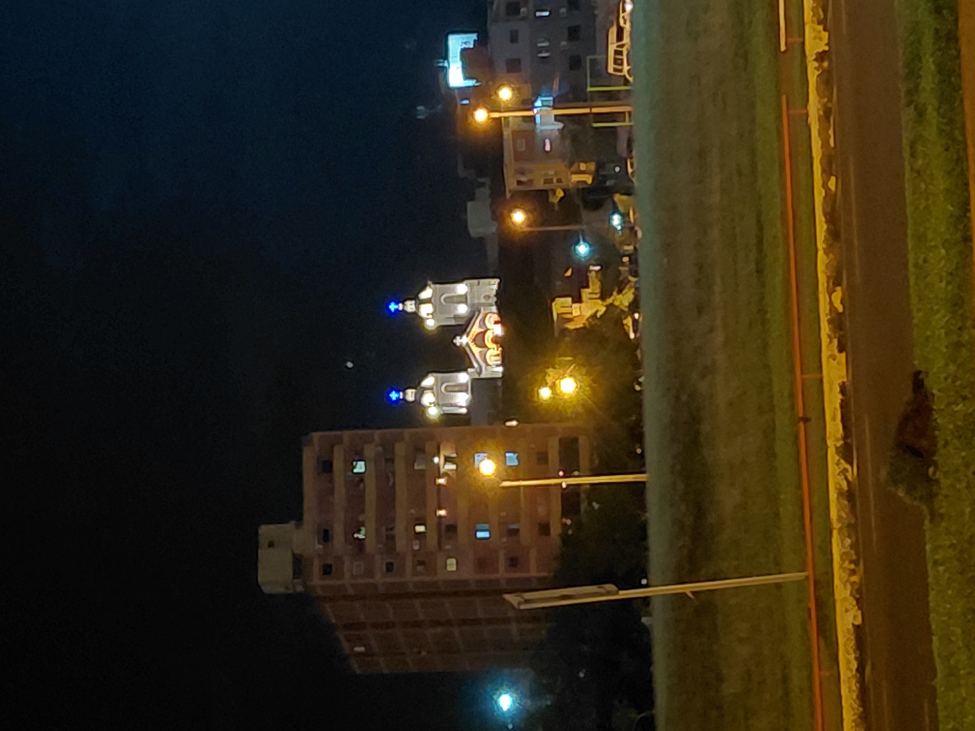 The image captures a nighttime view of a city in Encarnacion, Paraguay. In the background, a church with illuminated blue crosses on its domes stands out against the dark sky. Adjacent to the church, several buildings are visible, including a tall, multi-story brick building with lit windows. The foreground features a grassy slope, a road, and a yellow and orange striped barrier. Streetlights cast a warm glow on the scene, and a few vehicles are present, including a white van on the right side of the image. The overall atmosphere is urban and nocturnal.