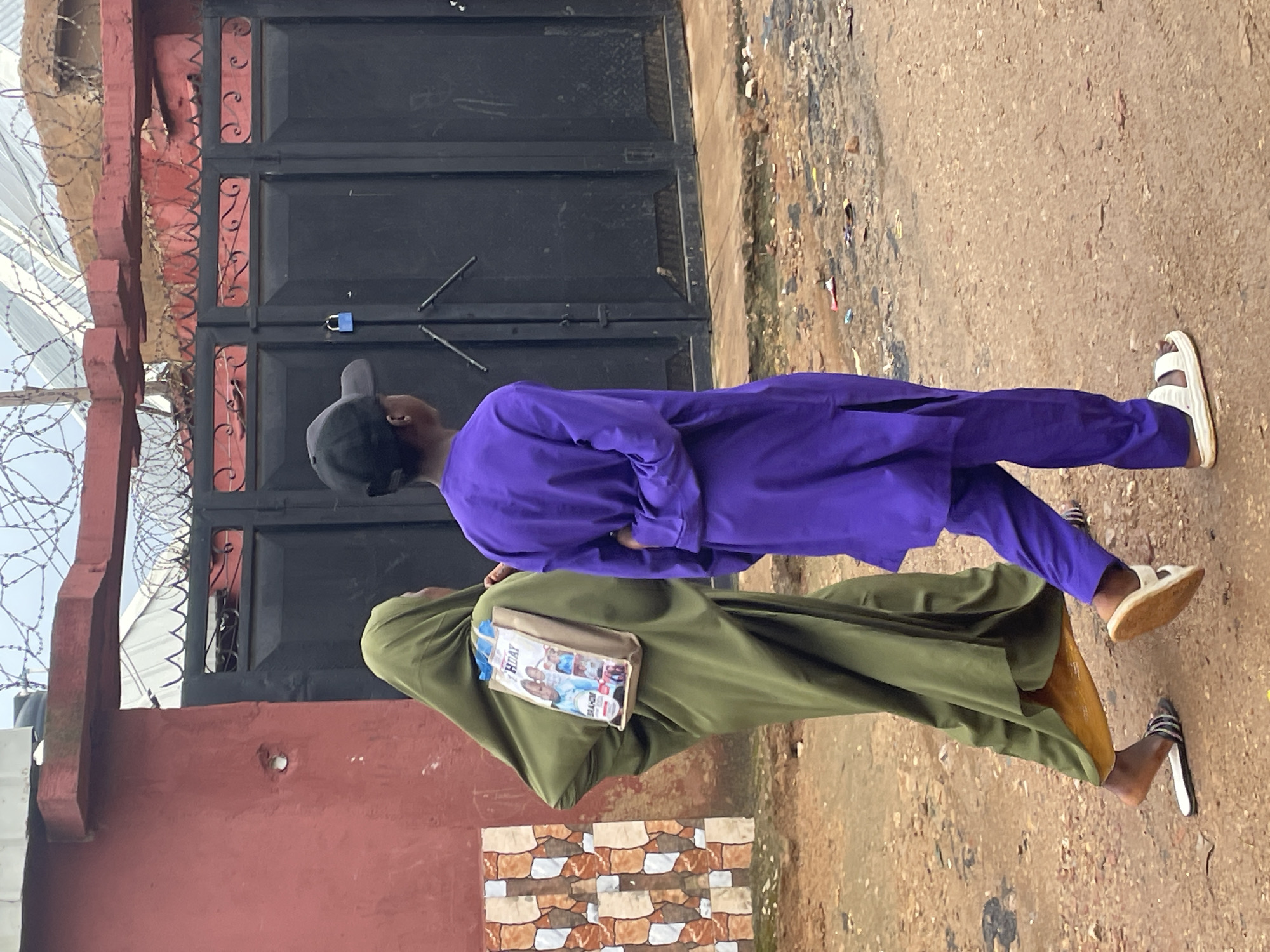In Gura Top, Nigeria, two individuals are walking on a dirt path in front of a large black metal gate topped with barbed wire. The person on the left, wearing a green hijab and a long green garment, carries a beige bag over their shoulder. The bag has images of children on it and text that appears to say "BIRTHDAY" and "BRAHIM". The person on the right, wearing a purple outfit and a black baseball cap, has their hands clasped behind their back. The gate is secured with a blue padlock. The ground is uneven and littered with some debris. A red wall with decorative tiling is visible to the left of the gate.
