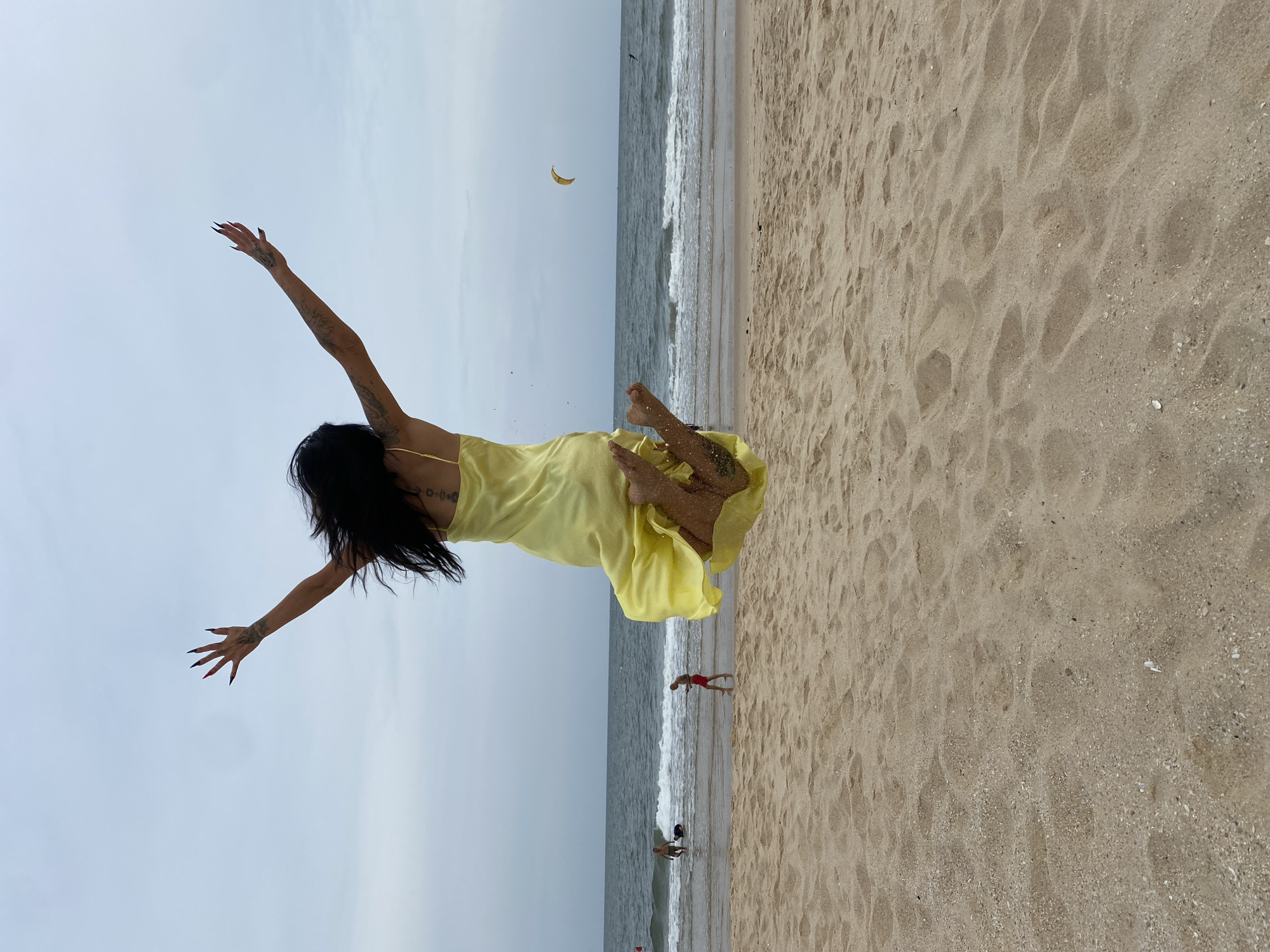 A woman in a yellow dress is suspended horizontally in the air on a beach. She has black hair and tattoos on her arm and back. Her arms are outstretched. There is sand on her feet. The beach has wet and dry sand, and the ocean is visible in the background. Two figures are partially submerged in the ocean's edge, and a third figure is wading in the water. A yellow object is in the sky. The location is Phu Thuy Ward, Vietnam.