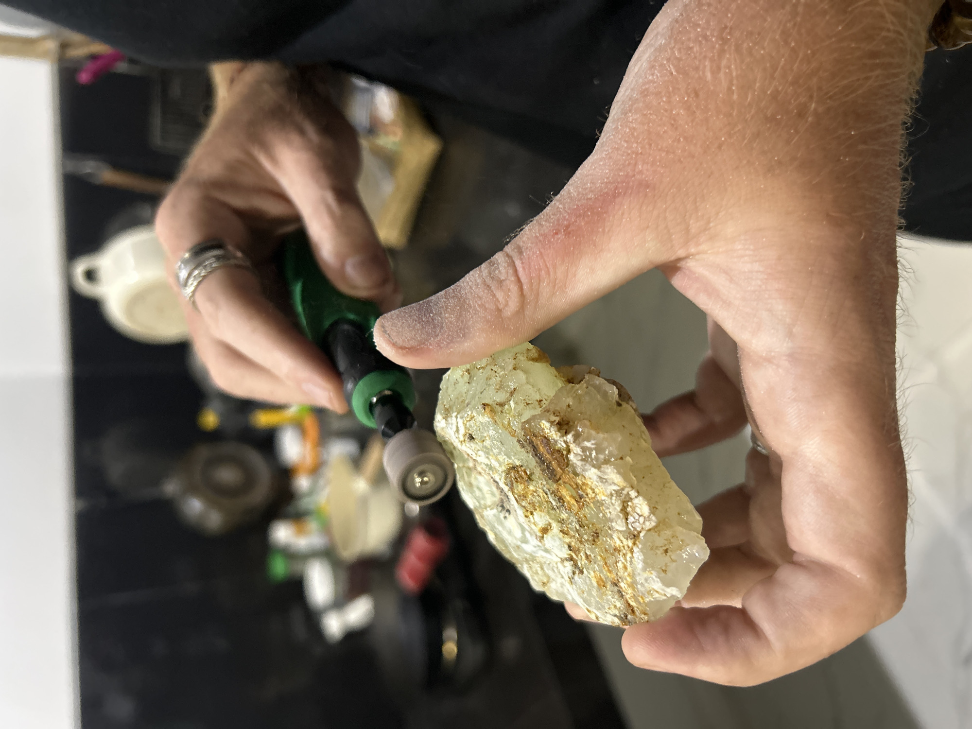The image shows a close-up of a person's hands working on a rough, translucent mineral specimen, likely a gemstone. The person is wearing rings on their left hand. In their right hand, they are holding a small, green rotary tool with a polishing attachment, which is in contact with the mineral. The mineral has a yellowish-white hue with streaks of brown and appears to be partly polished and partly in its raw state. The background is blurred, but suggests a workshop or crafting area with various tools and materials visible, including what looks like other mineral samples and containers. The overall lighting suggests an indoor setting, possibly during the day. The presence of dust on the hands and the tool indicates an active crafting process. The scene conveys a sense of focused activity and craftsmanship. The location is identified as Sukawati, Indonesia, which is known for its art markets and traditional crafts.